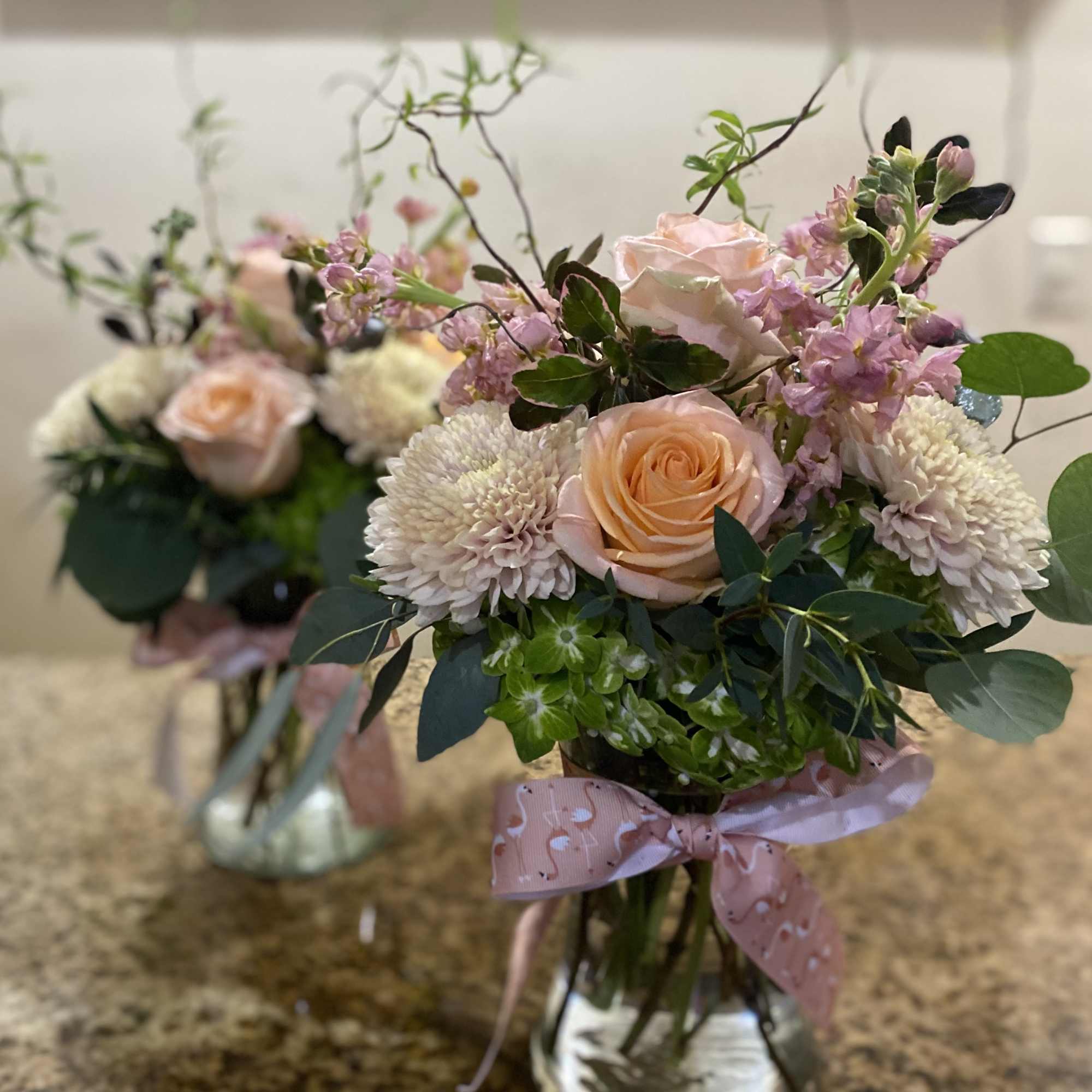 Two pastel peach rose and white mum arrangements in glass vases with pink bows on a granite countertop