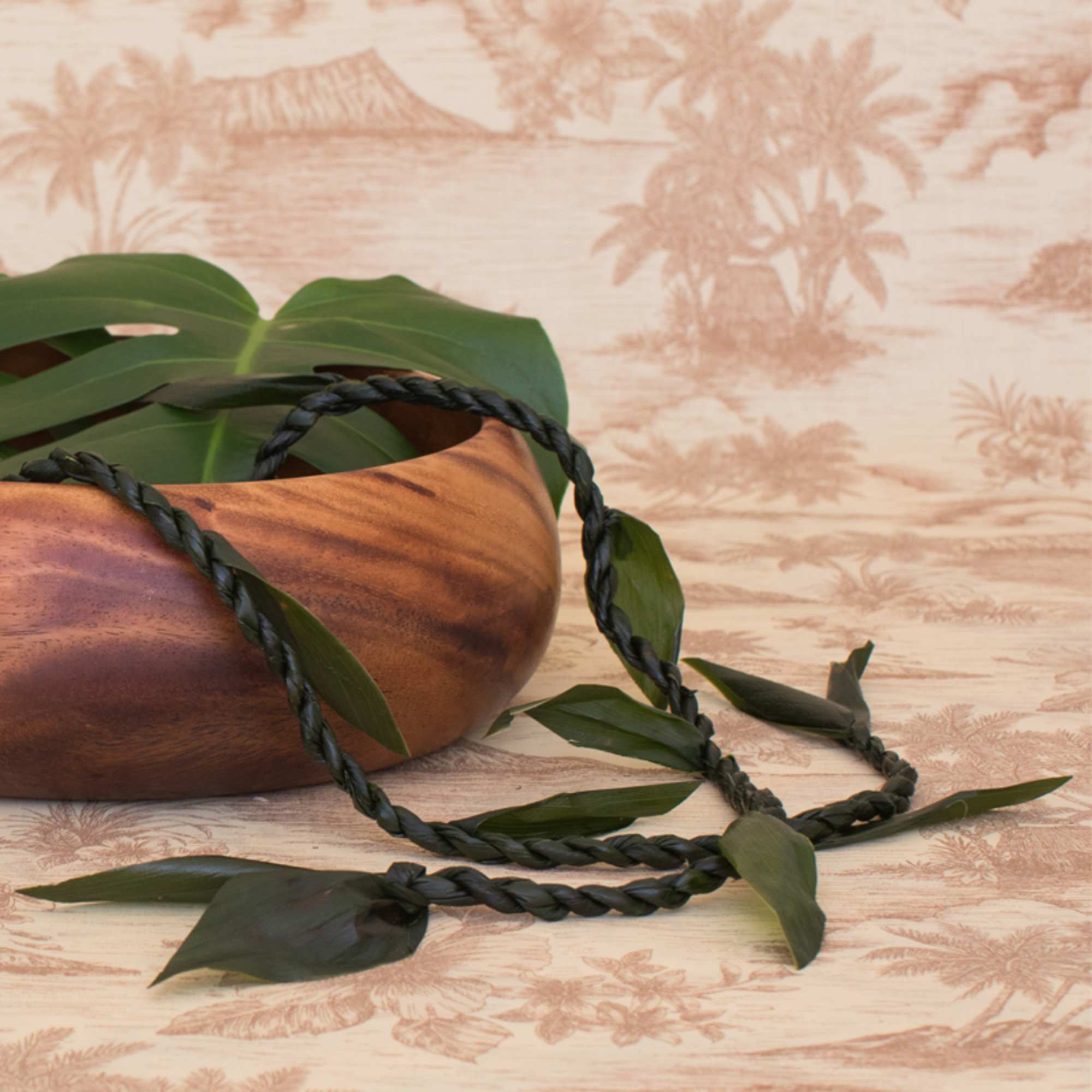 Braided dark green leaf lei draped over a wooden bowl with tropical backdrop