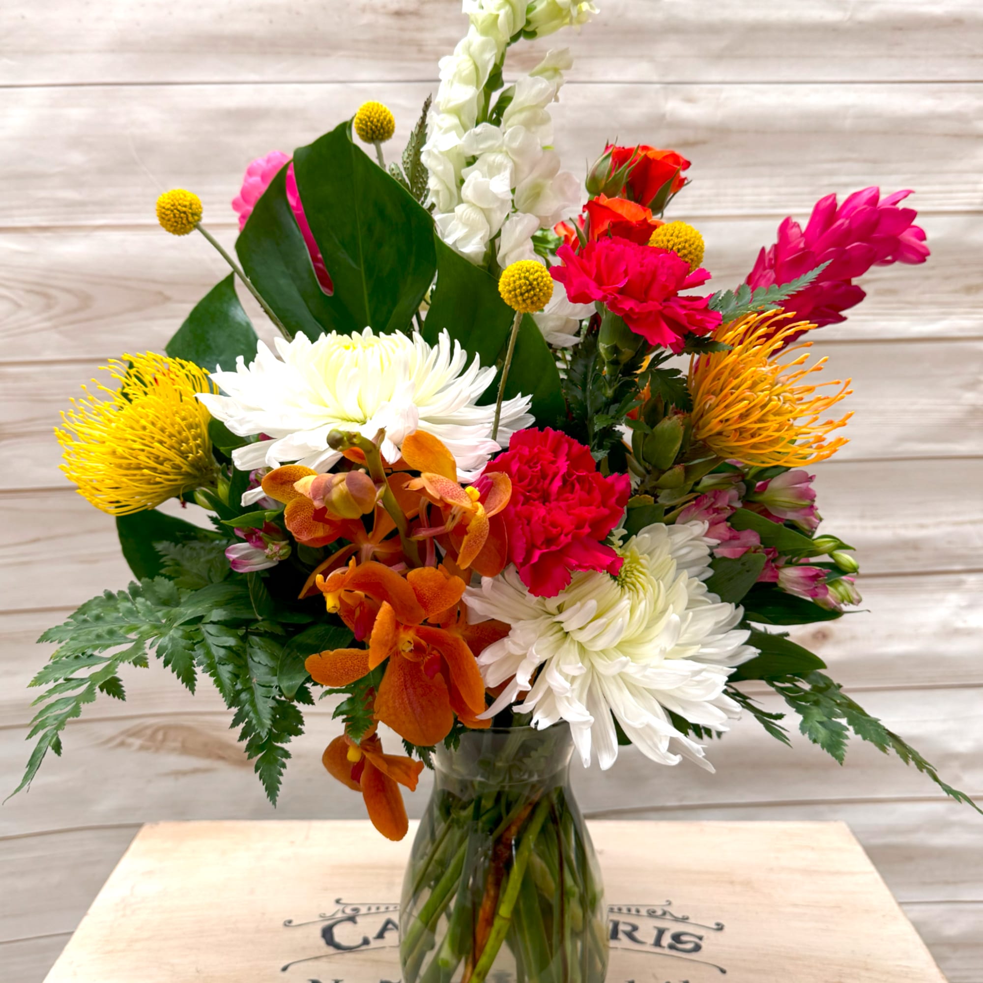 Tall mixed arrangement with white mums, orange orchids, pink carnations, and yellow protea in a clear glass vase