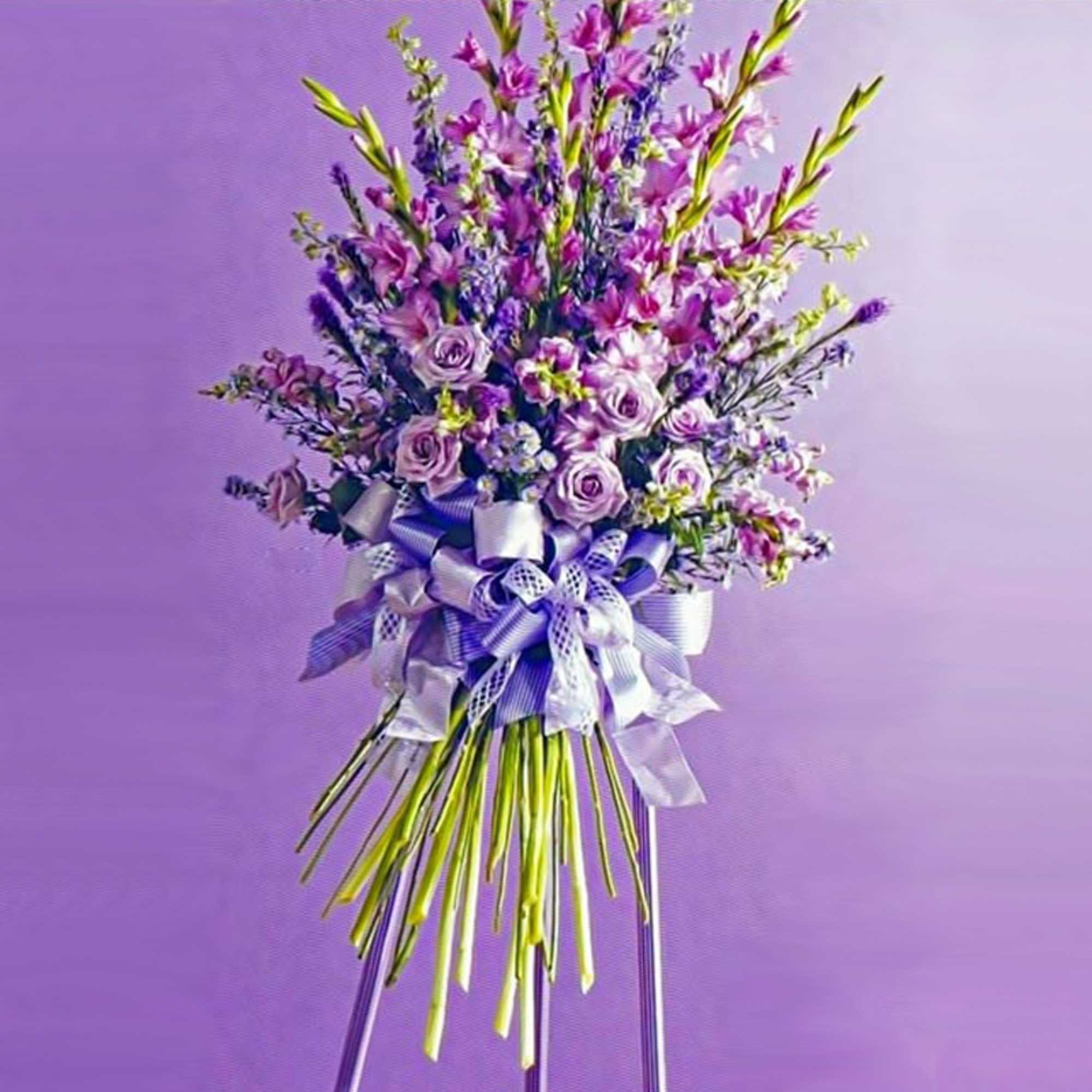 Tall standing arrangement of purple and pink flowers with large lavender ribbons on a tripod stand