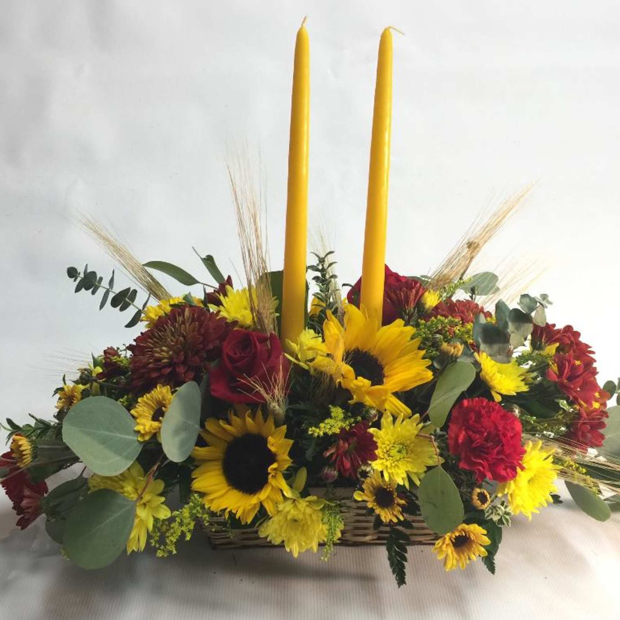 Sunflower and red flower arrangement with two tall yellow candles in a basket