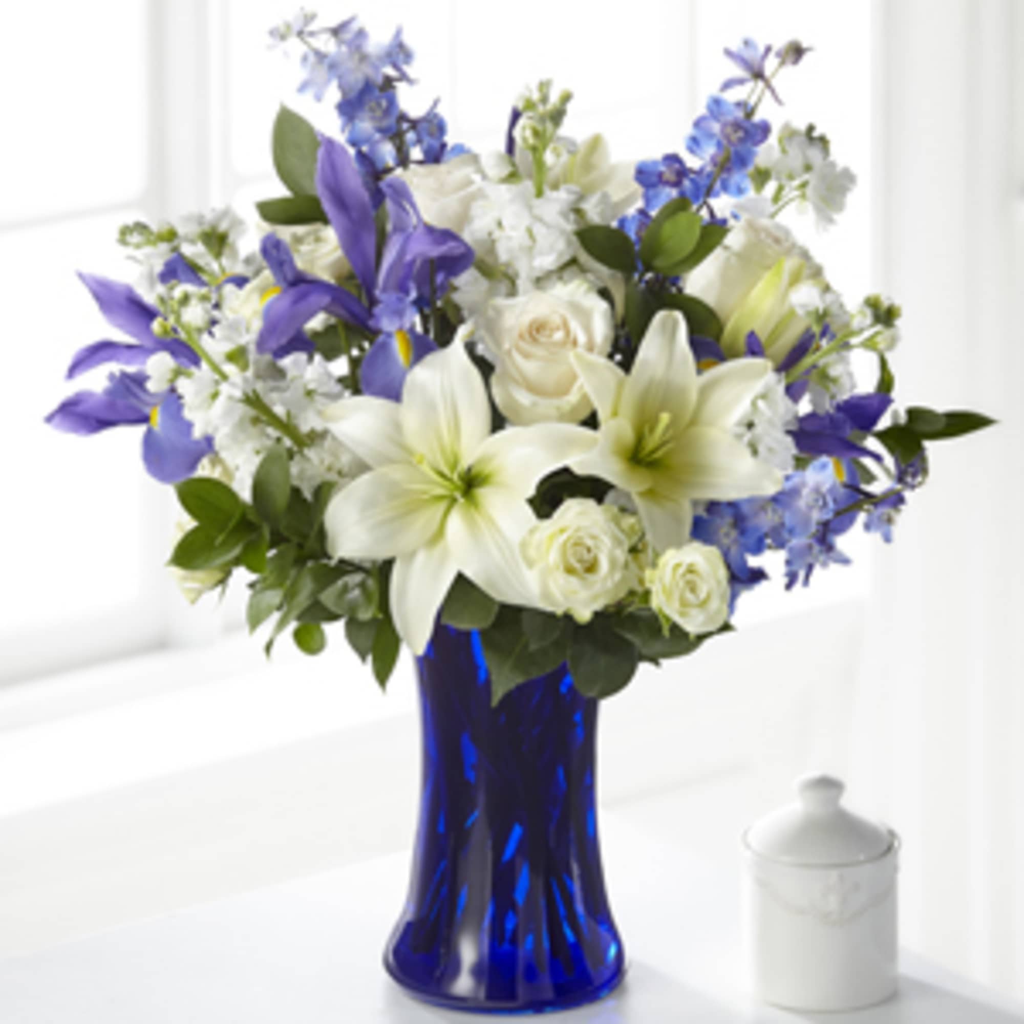 White lilies, roses, and blue flowers arranged in a cobalt blue glass vase