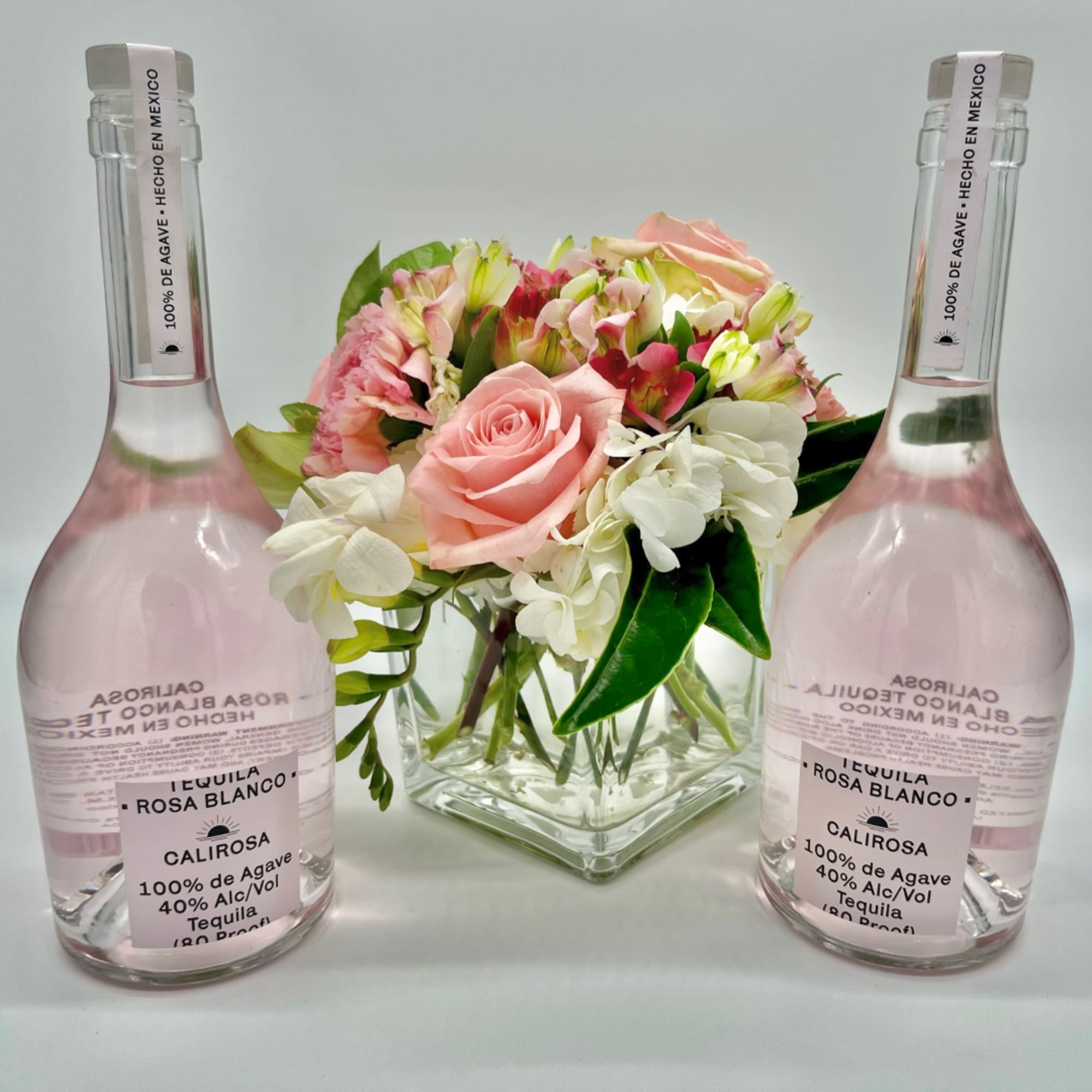 Pink and white flower arrangement in a square glass vase, flanked by two tequila bottles