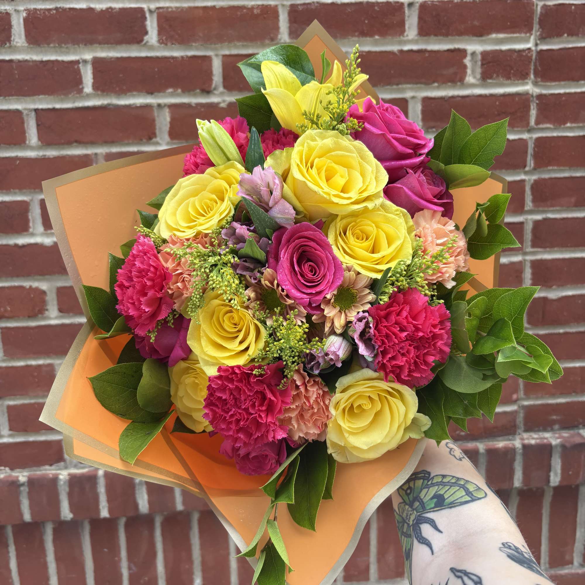 Handheld bouquet of yellow and pink roses with carnations in orange wrap