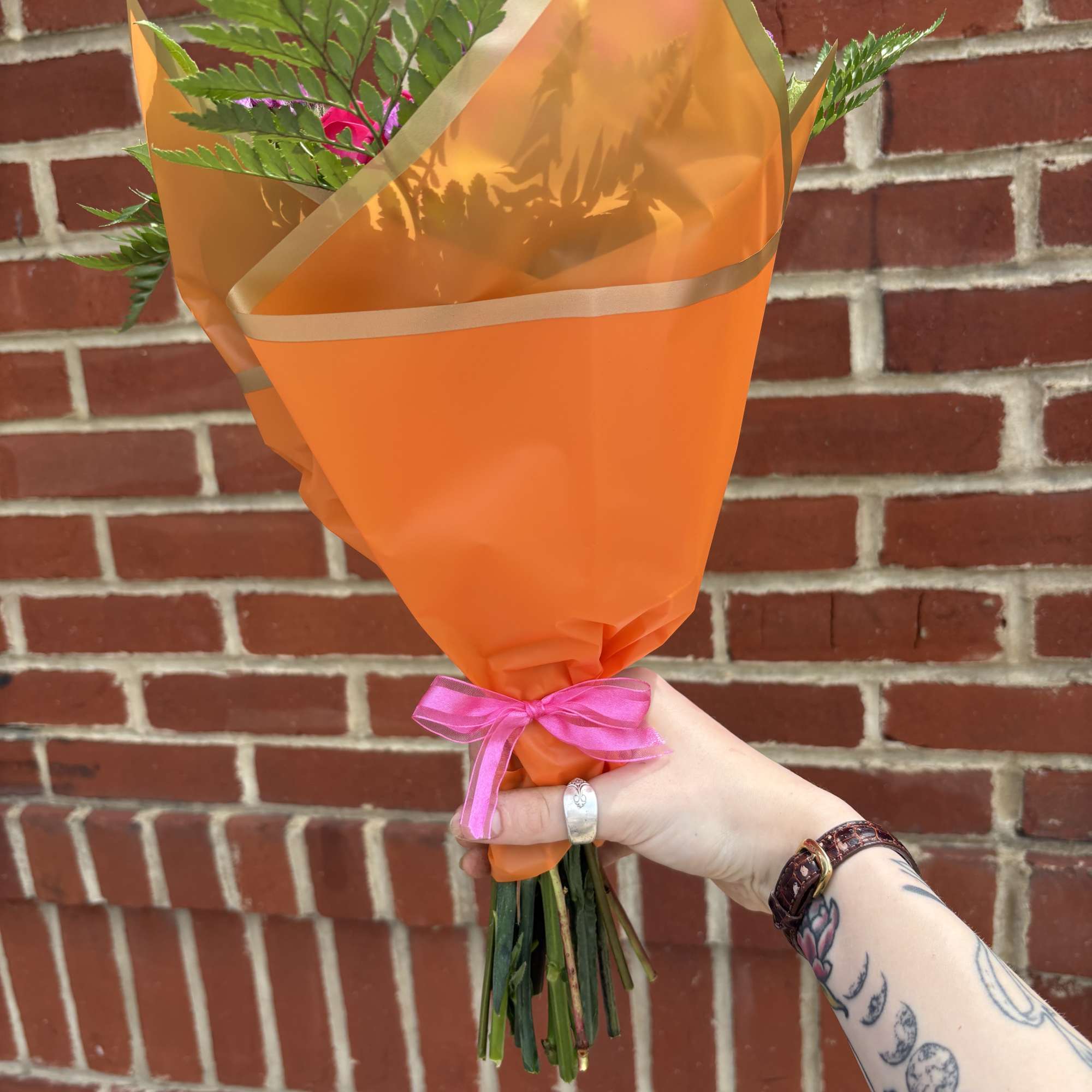 Hand holding an orange-wrapped bouquet with pink ribbon and fern foliage