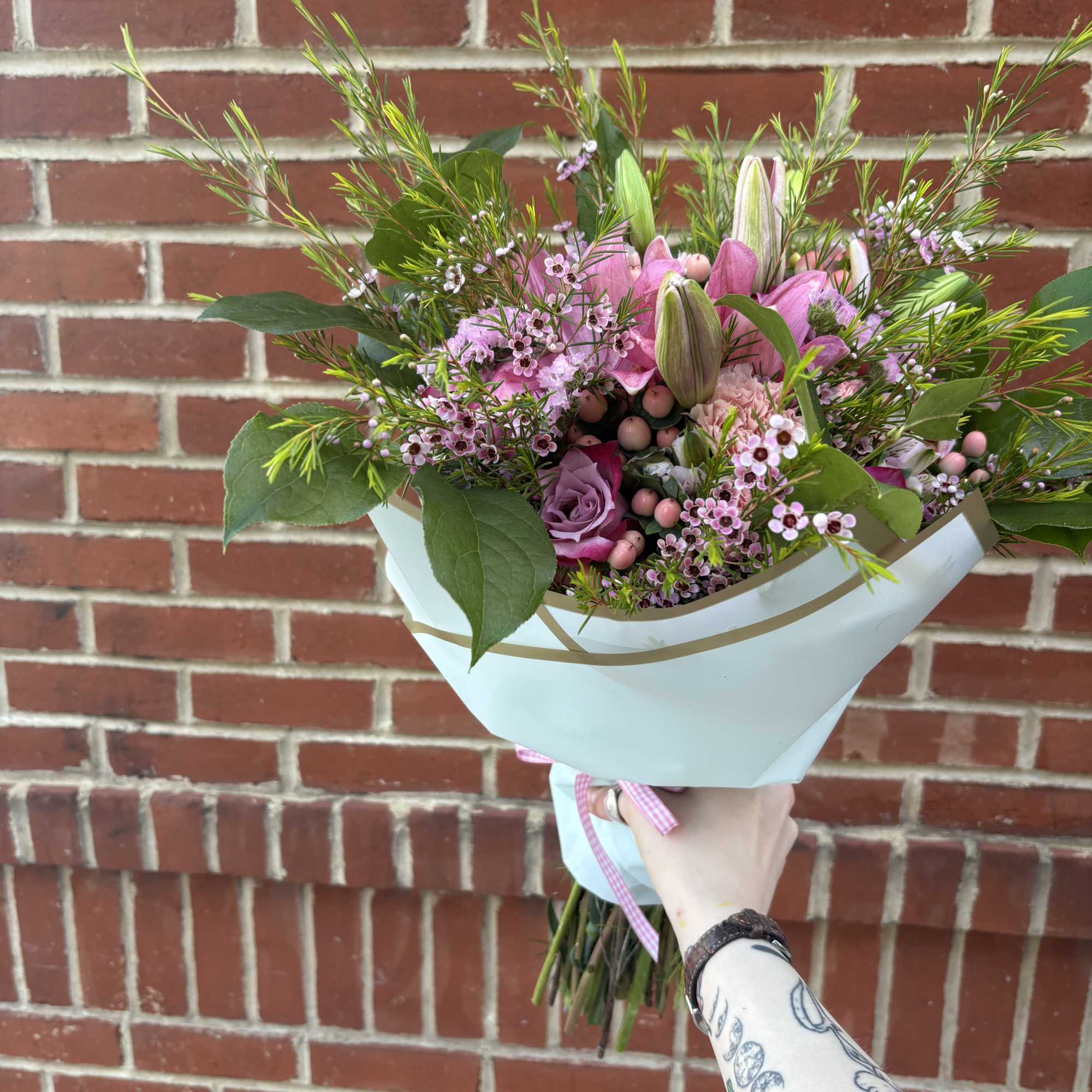 Handheld bouquet of pink flowers with white wrapping against a brick wall
