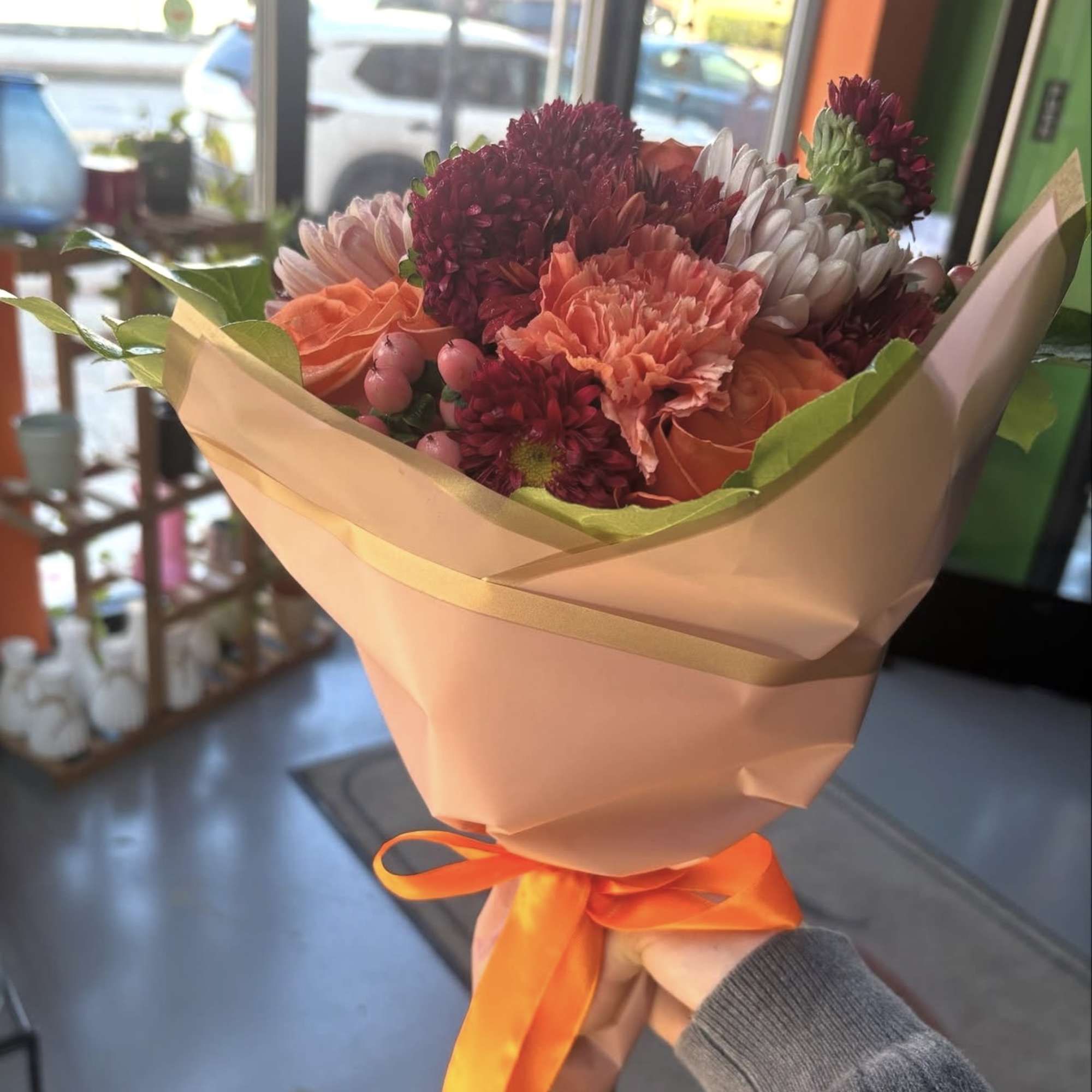 Handheld bouquet of orange and burgundy flowers wrapped in peach paper