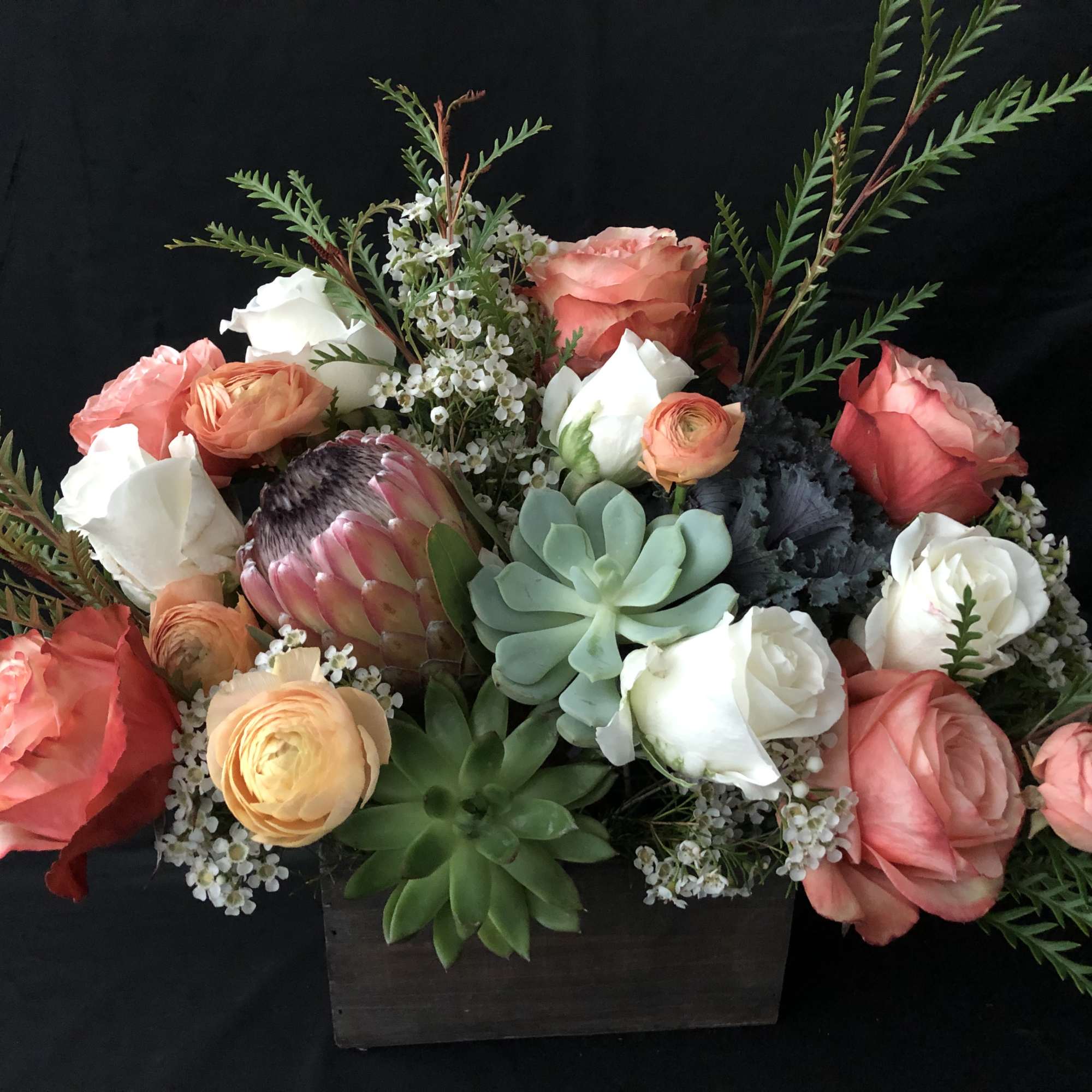 Low wooden box arrangement with peach and white roses, succulents, ranunculus, and a central pink protea.