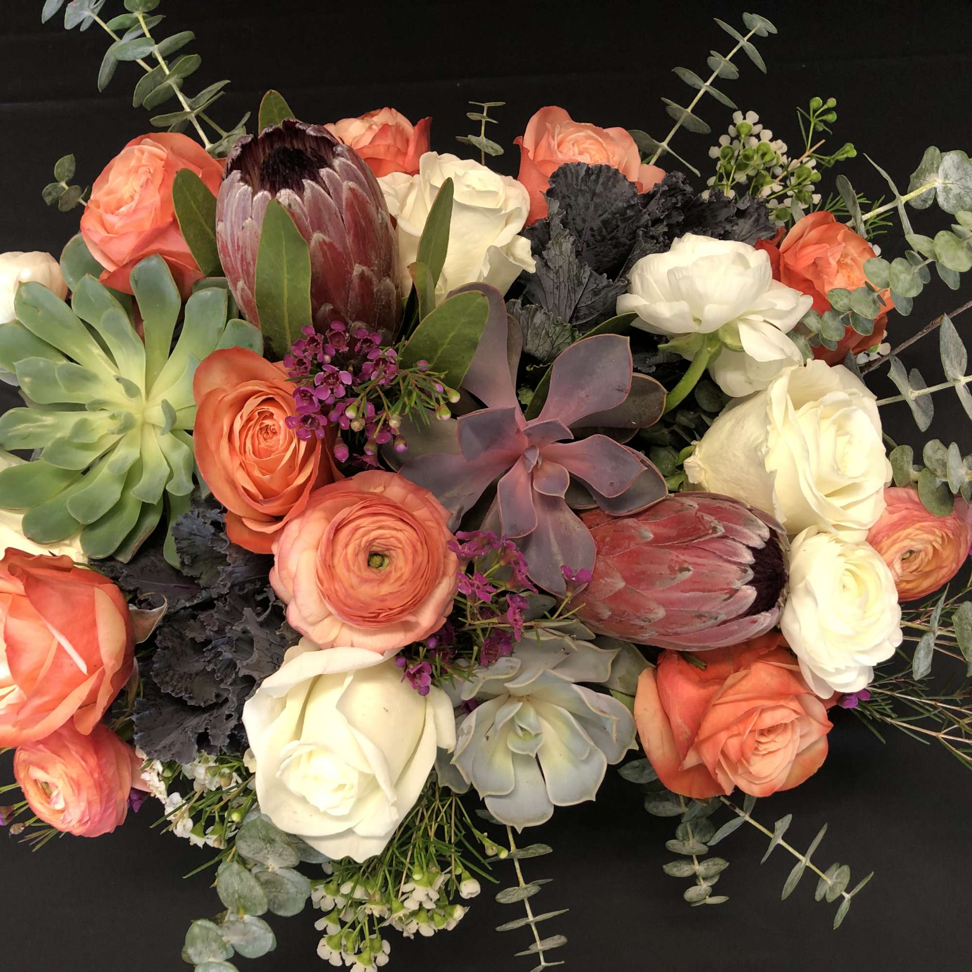 Low arrangement of orange and white roses with succulents and protea in a compact centerpiece.