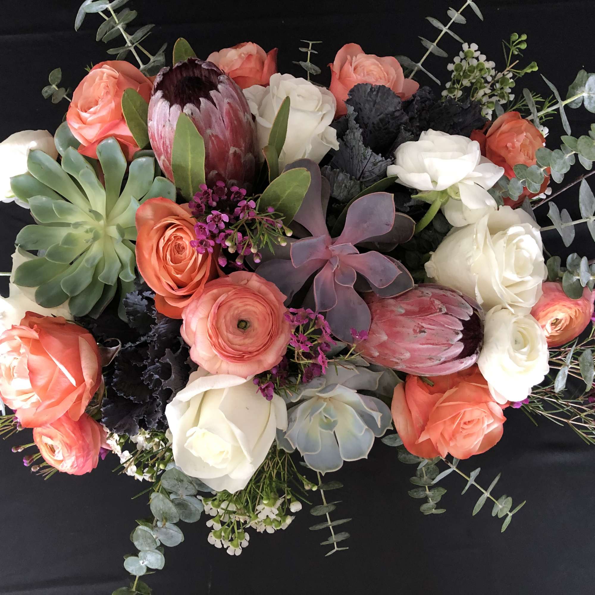 Low arrangement of peach and white roses, succulents, and pink protea on a dark background