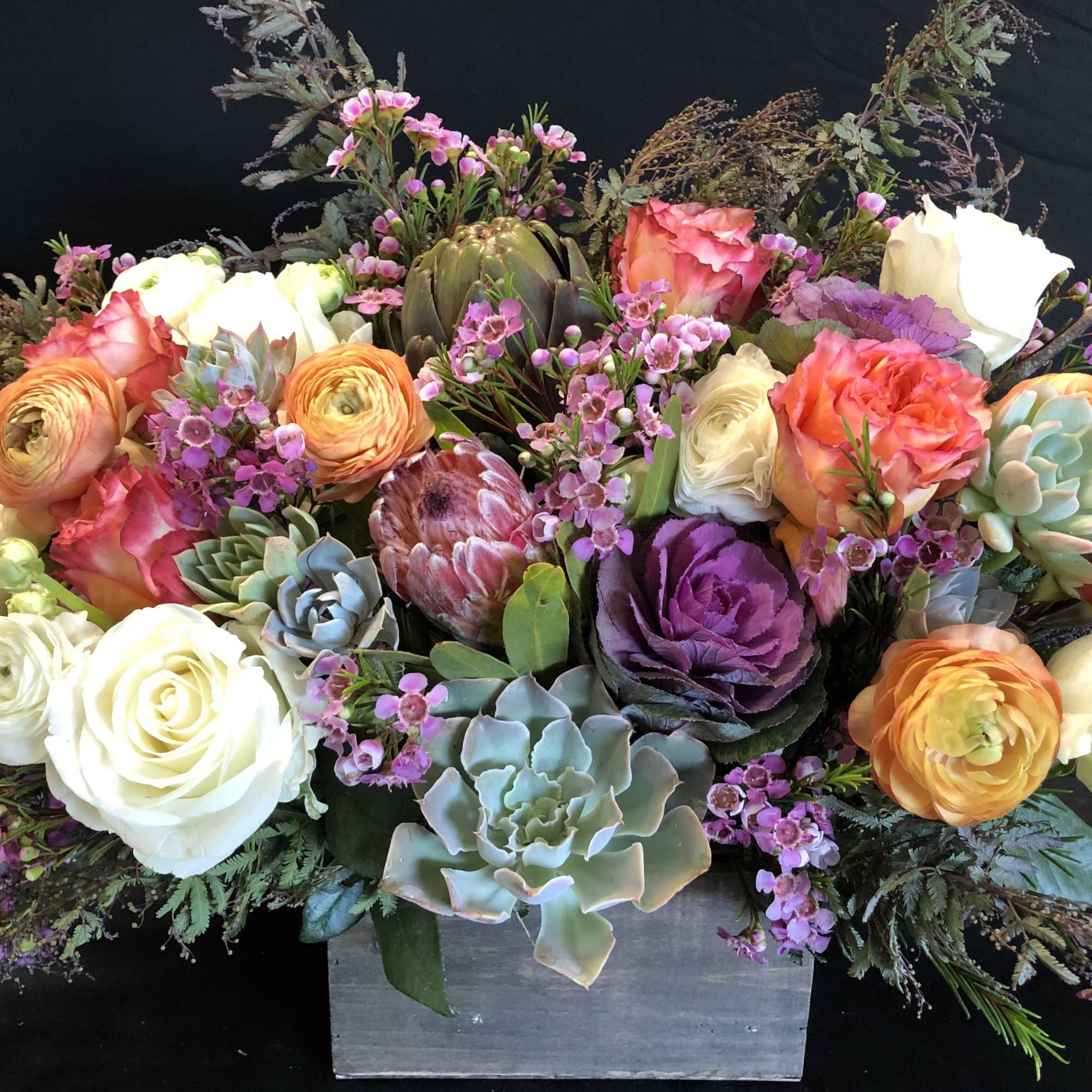 Lush arrangement of roses, ranunculus, succulents, and protea in a wooden box.