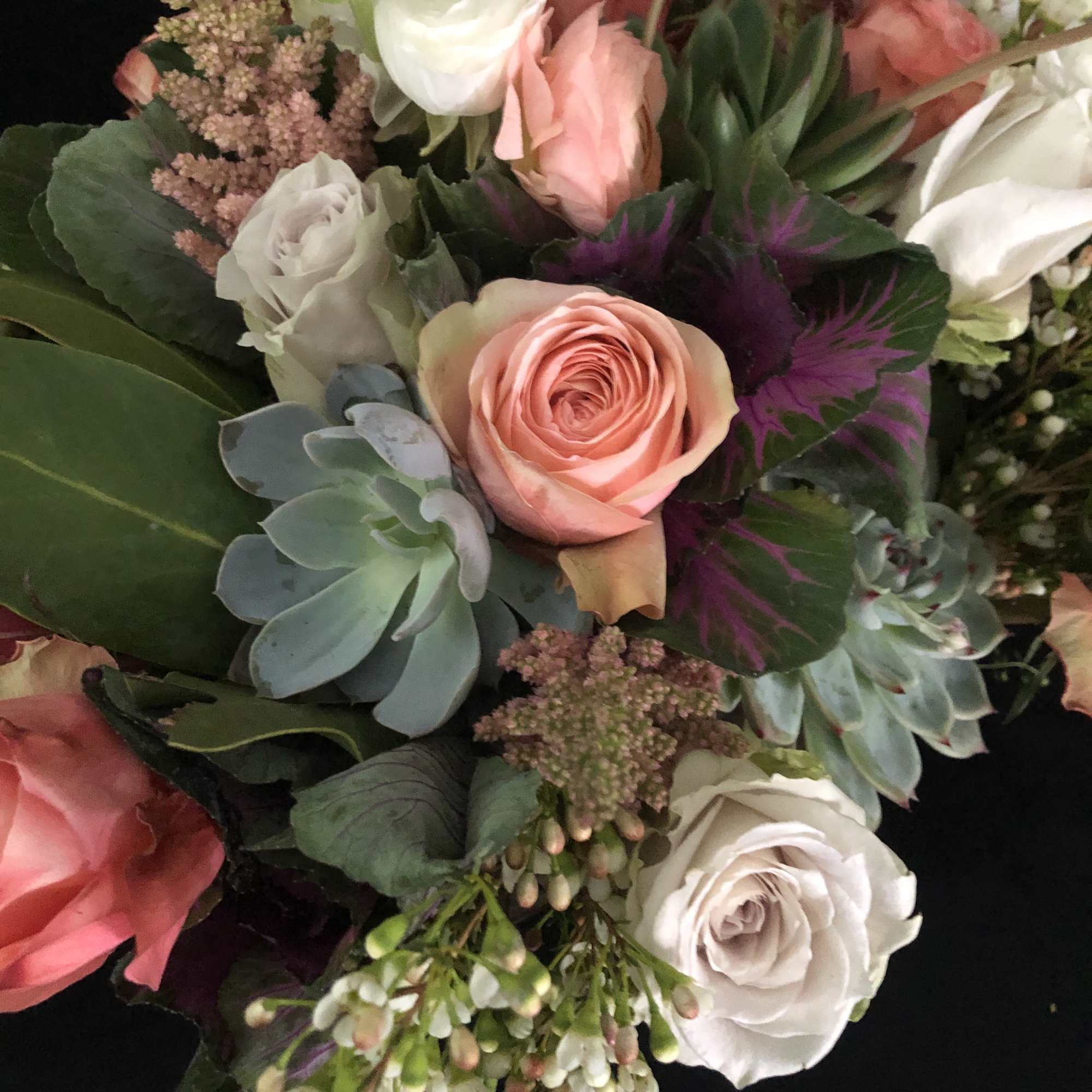Bouquet of peach and white roses, ranunculus, and succulents with purple accents on a dark background