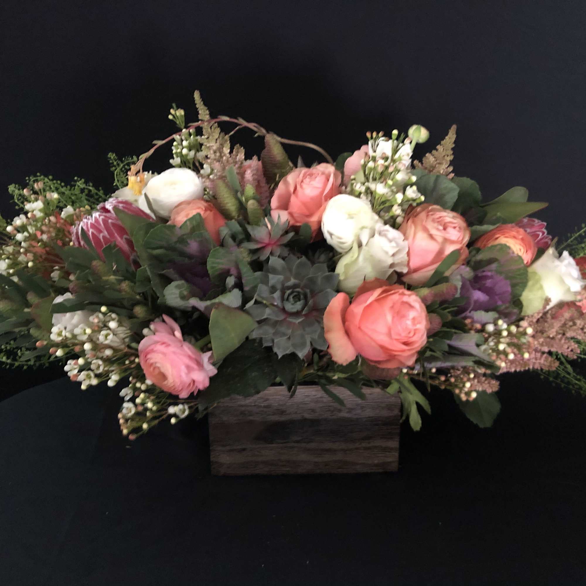 Low arrangement of peach and white roses with succulents and protea in a rectangular wooden box.