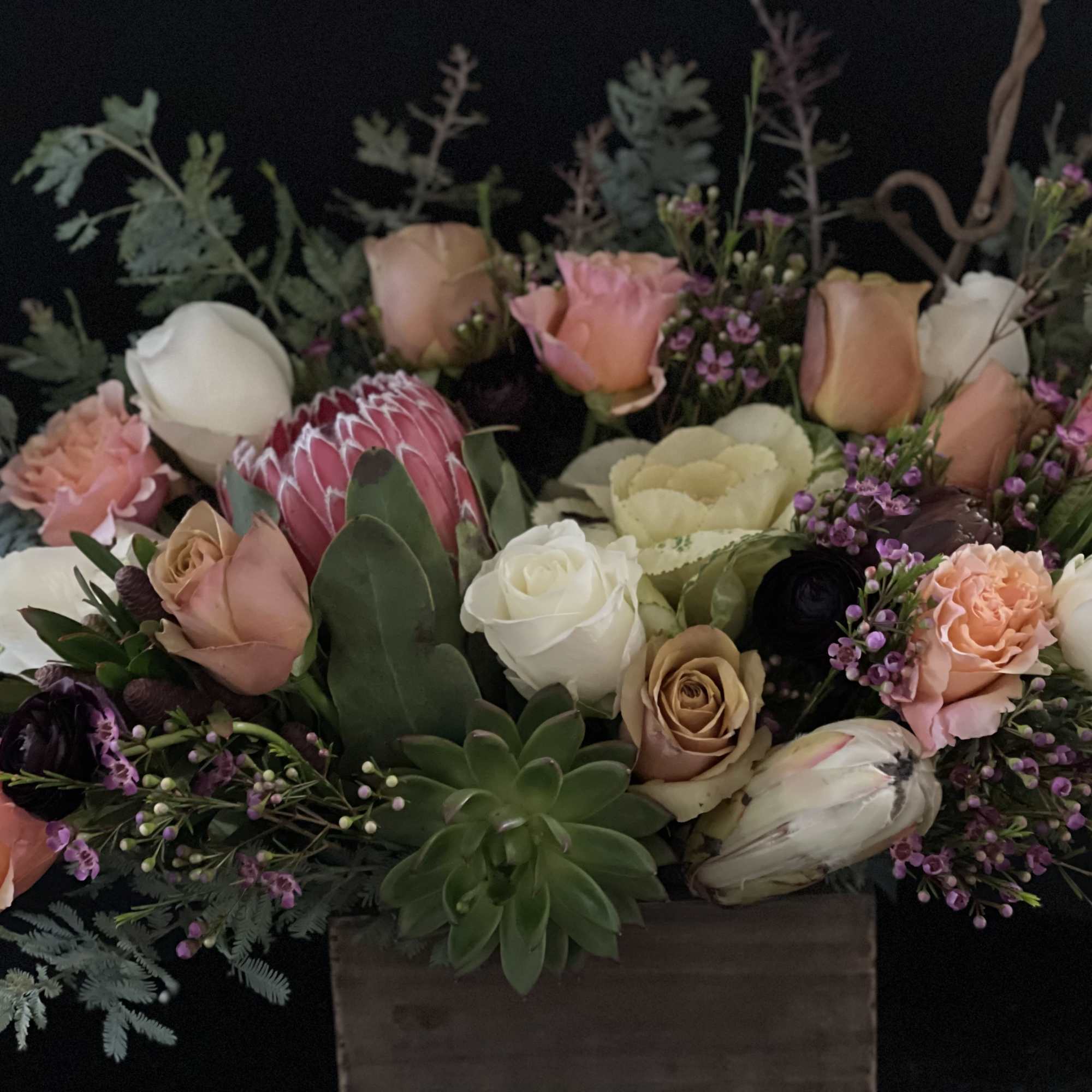 Low wooden box arrangement with peach and white roses, protea, succulents, and mixed blooms.