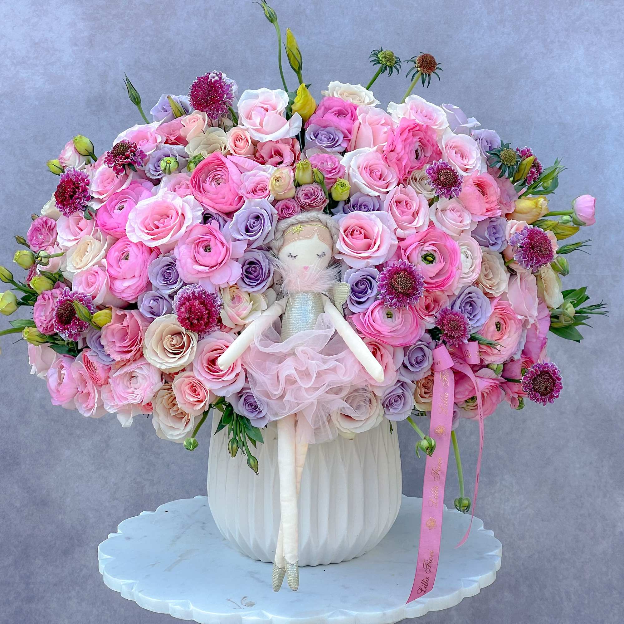 Large pink and lavender rose arrangement with a doll in a white vase