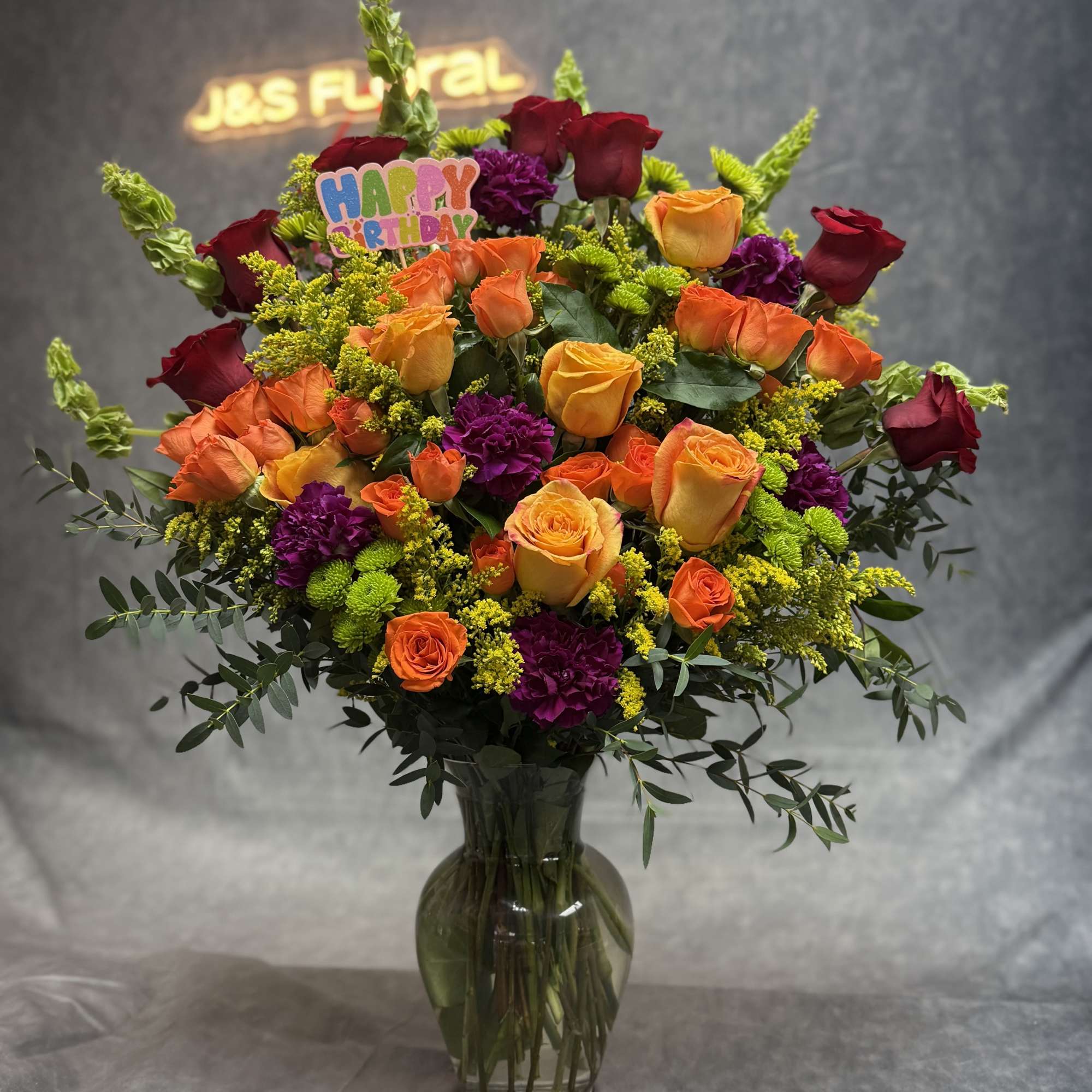Mixed orange and red roses in a glass vase with a birthday pick