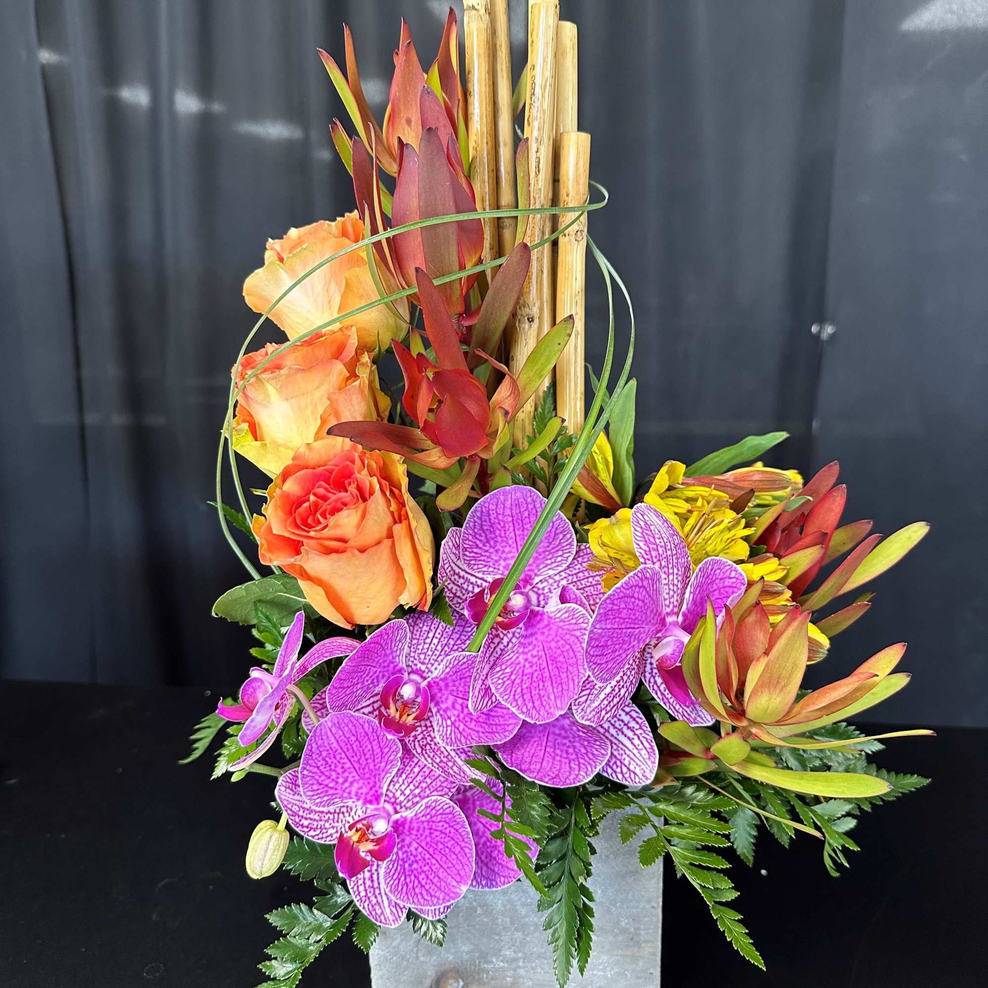 Orange roses and purple orchids in a square metal vase with bamboo accents