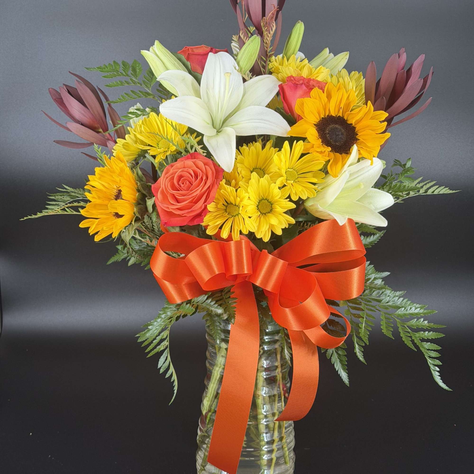 Mixed bouquet of lilies, sunflowers, roses, and yellow daisies in a glass vase with an orange ribbon