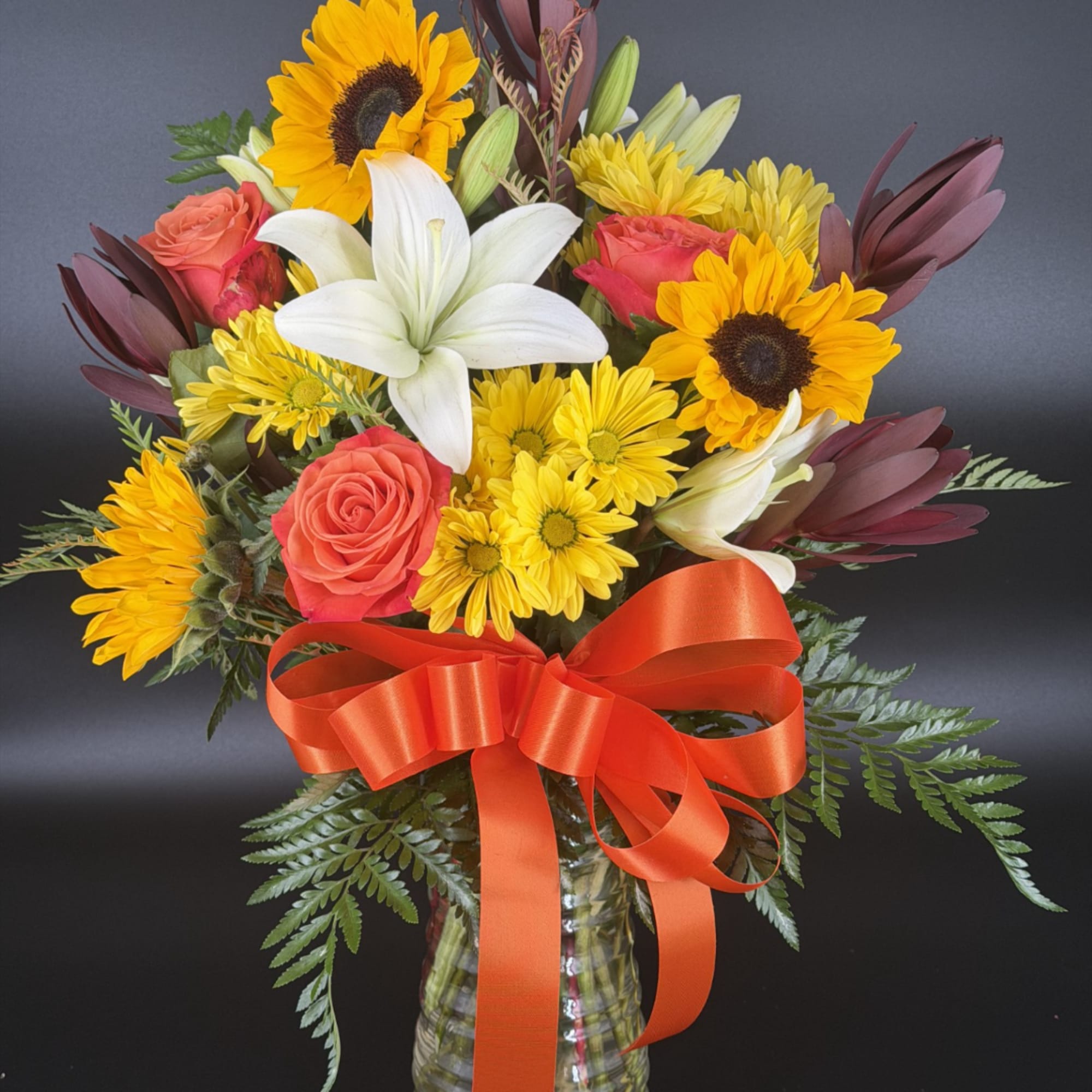 Bouquet of sunflowers, white lilies, roses, and yellow daisies in a glass vase with an orange ribbon