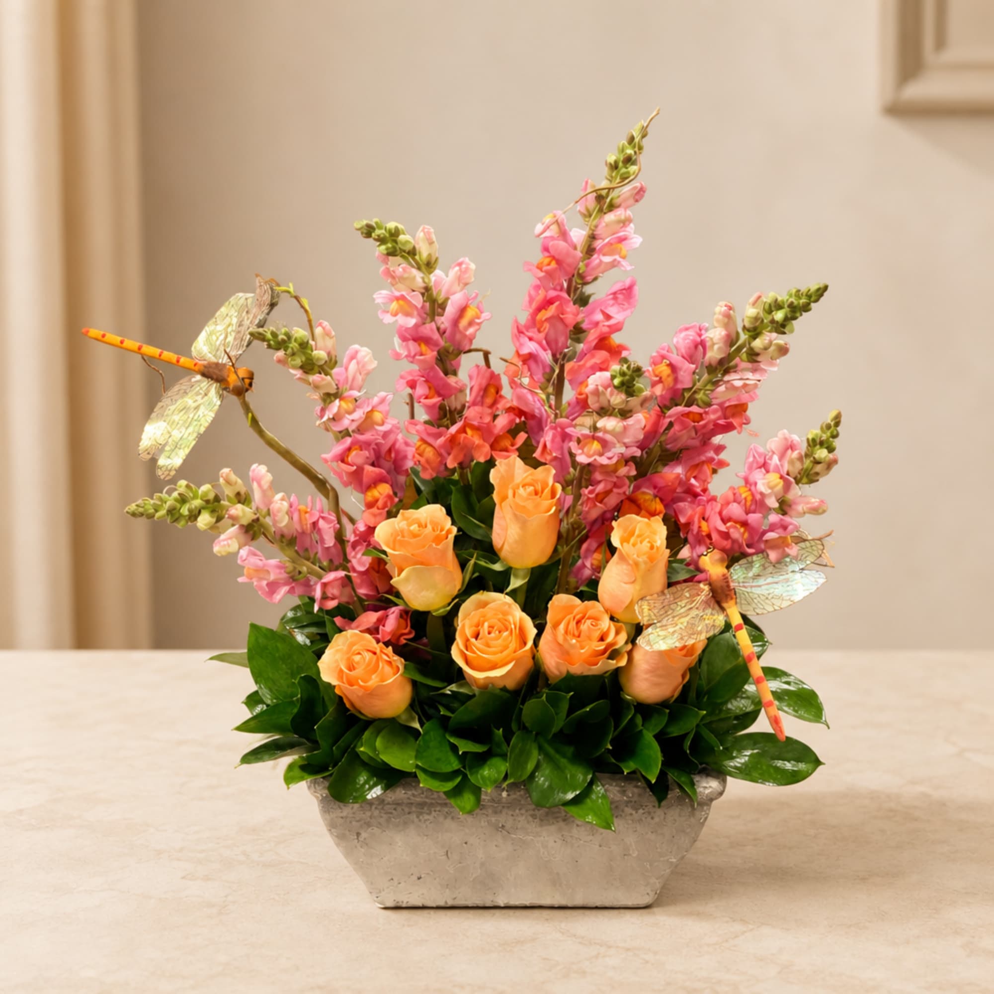 Orange roses and pink snapdragons in a rectangular vase with dragonfly picks