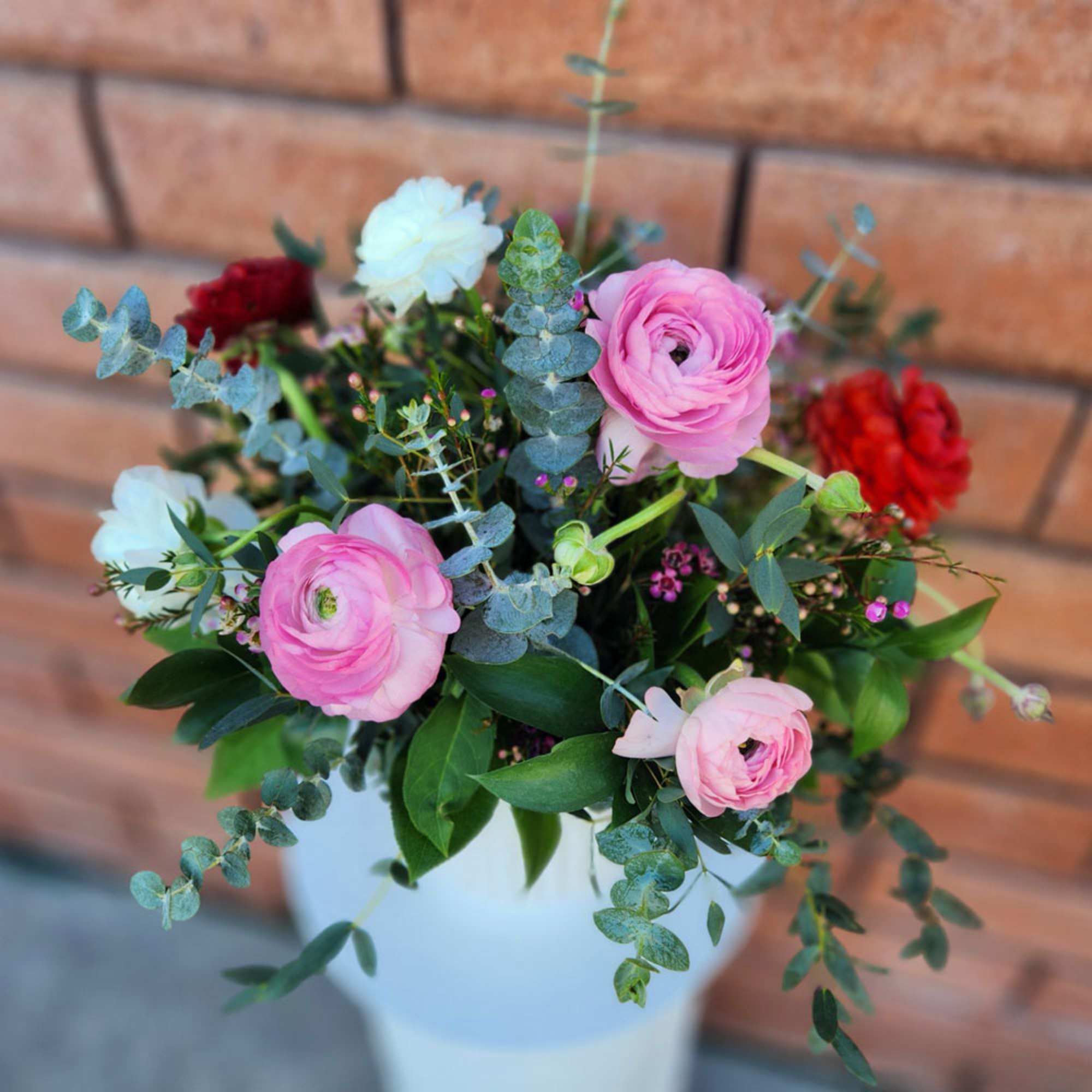 Arrangement of pink, white, and red ranunculus in a white vase against a brick wall