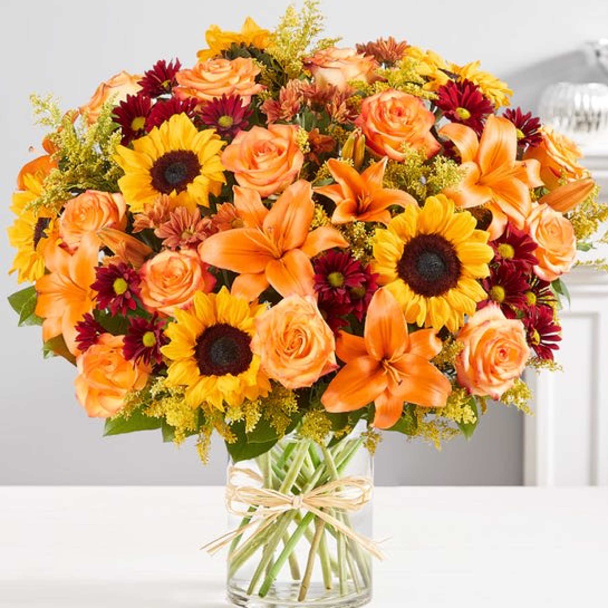 Bouquet of orange roses, sunflowers, lilies, and burgundy daisies in a clear glass vase