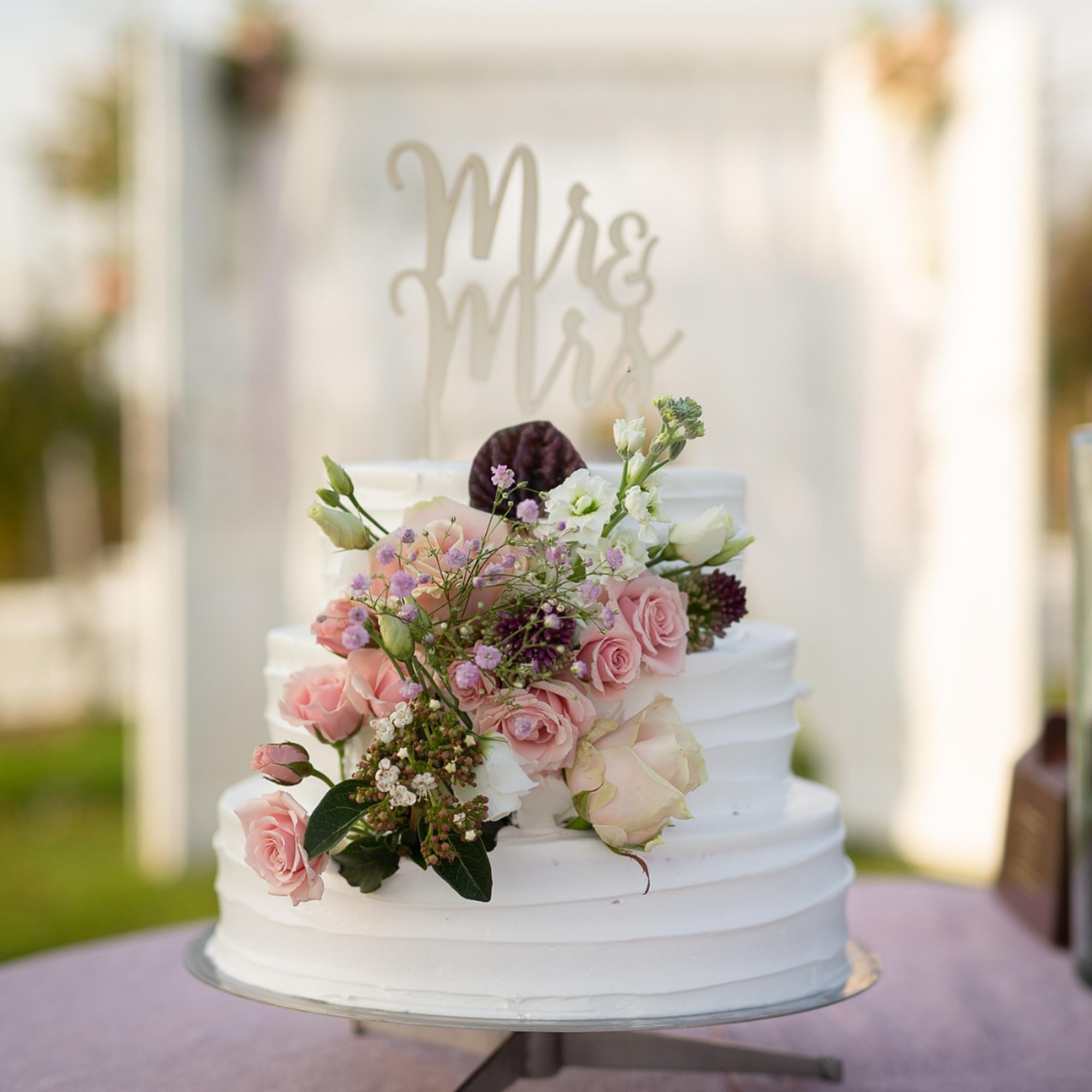 White tiered wedding cake topped with pink roses and small purple flowers