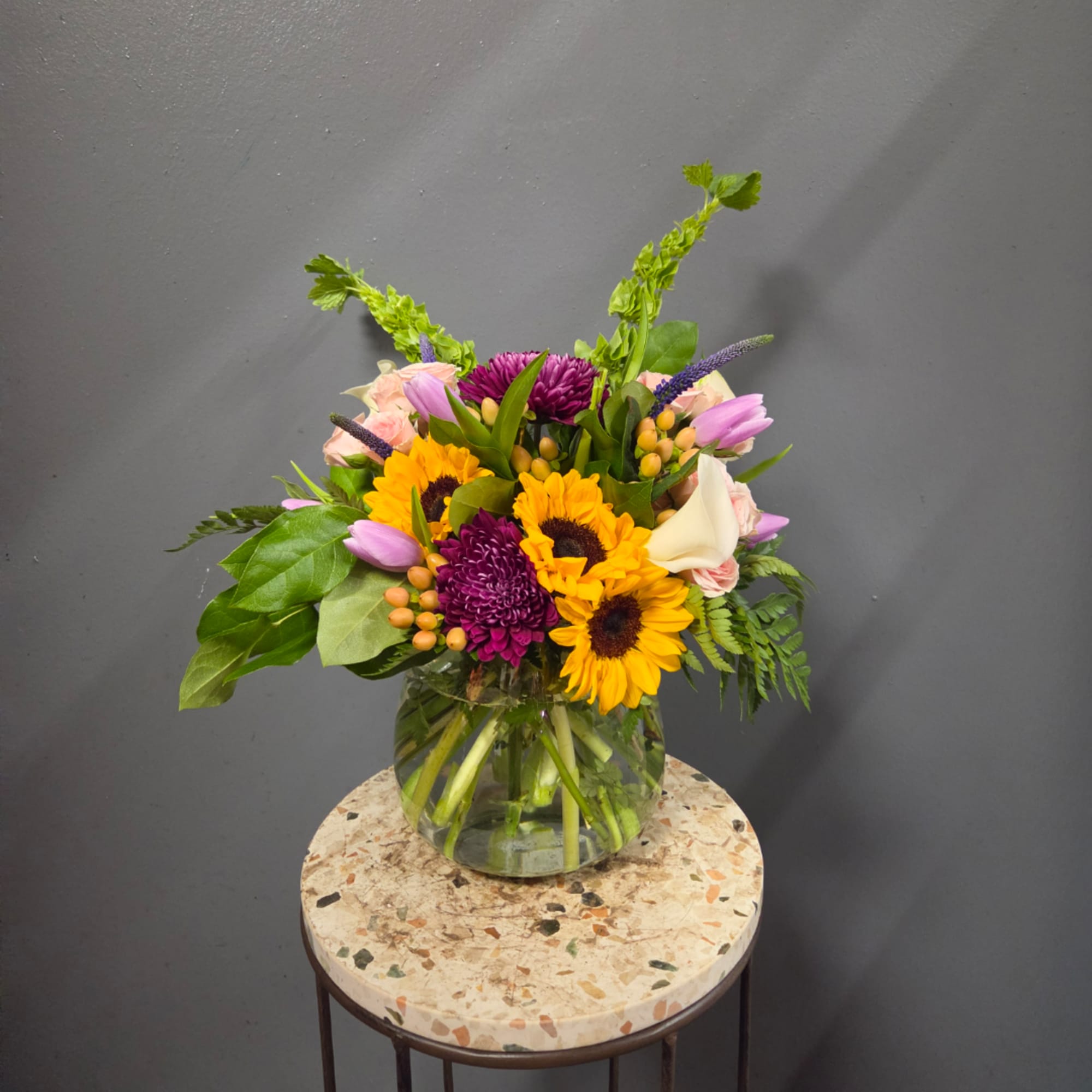 Mixed bouquet in a clear glass vase with sunflowers, purple blooms, and pink tulips