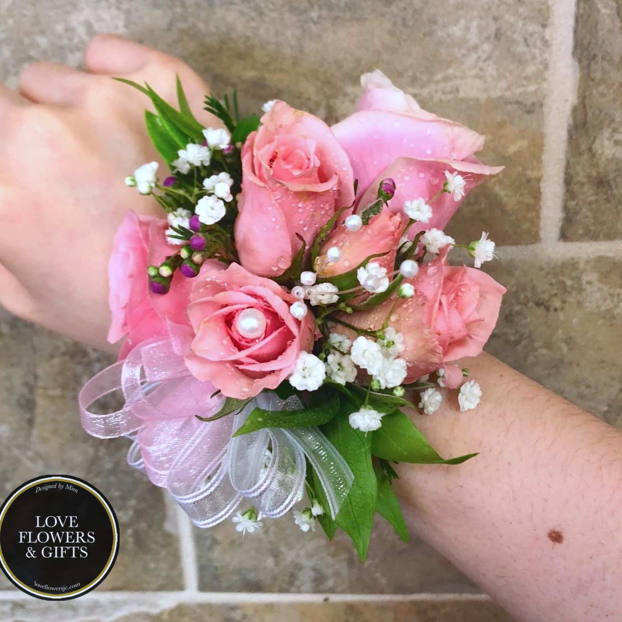 Pink rose wrist corsage with white filler flowers, pearls, and sheer ribbon on a wrist
