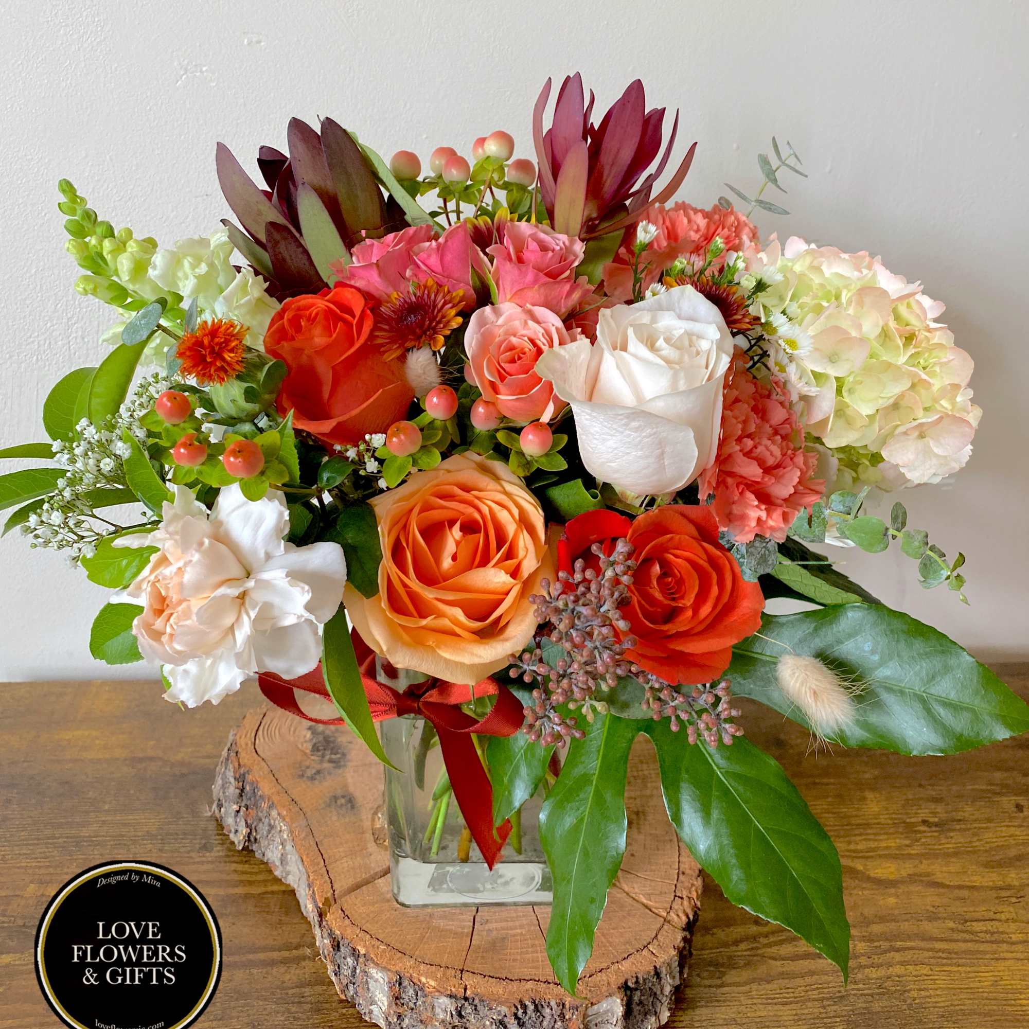 Mixed orange, white, and pink flower arrangement in a clear glass vase on a wood slice base
