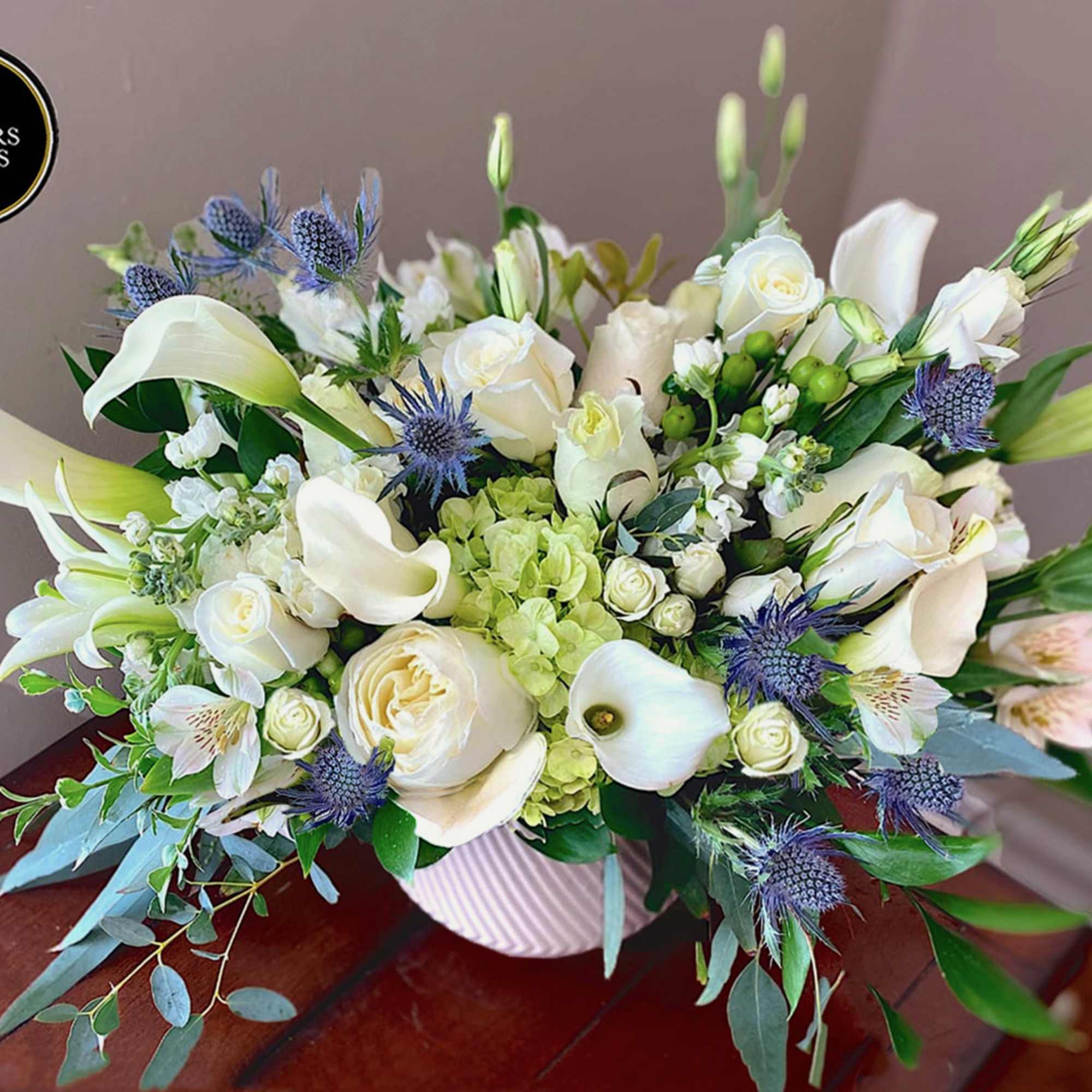 Low white and green arrangement of roses, calla lilies, and blue thistle in a white ribbed vase