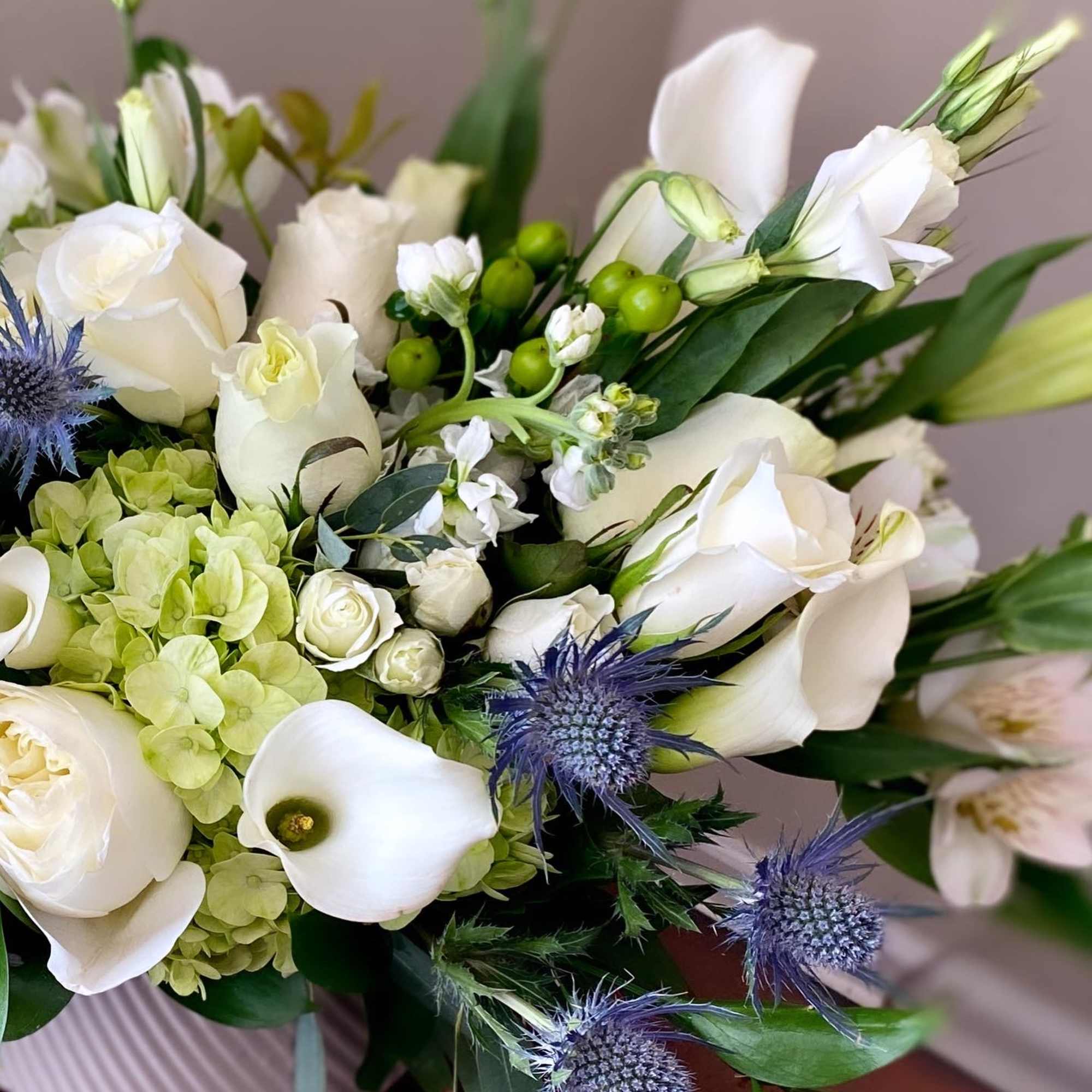 Low arrangement of white roses, calla lilies, hydrangeas and blue thistle in a white ceramic bowl