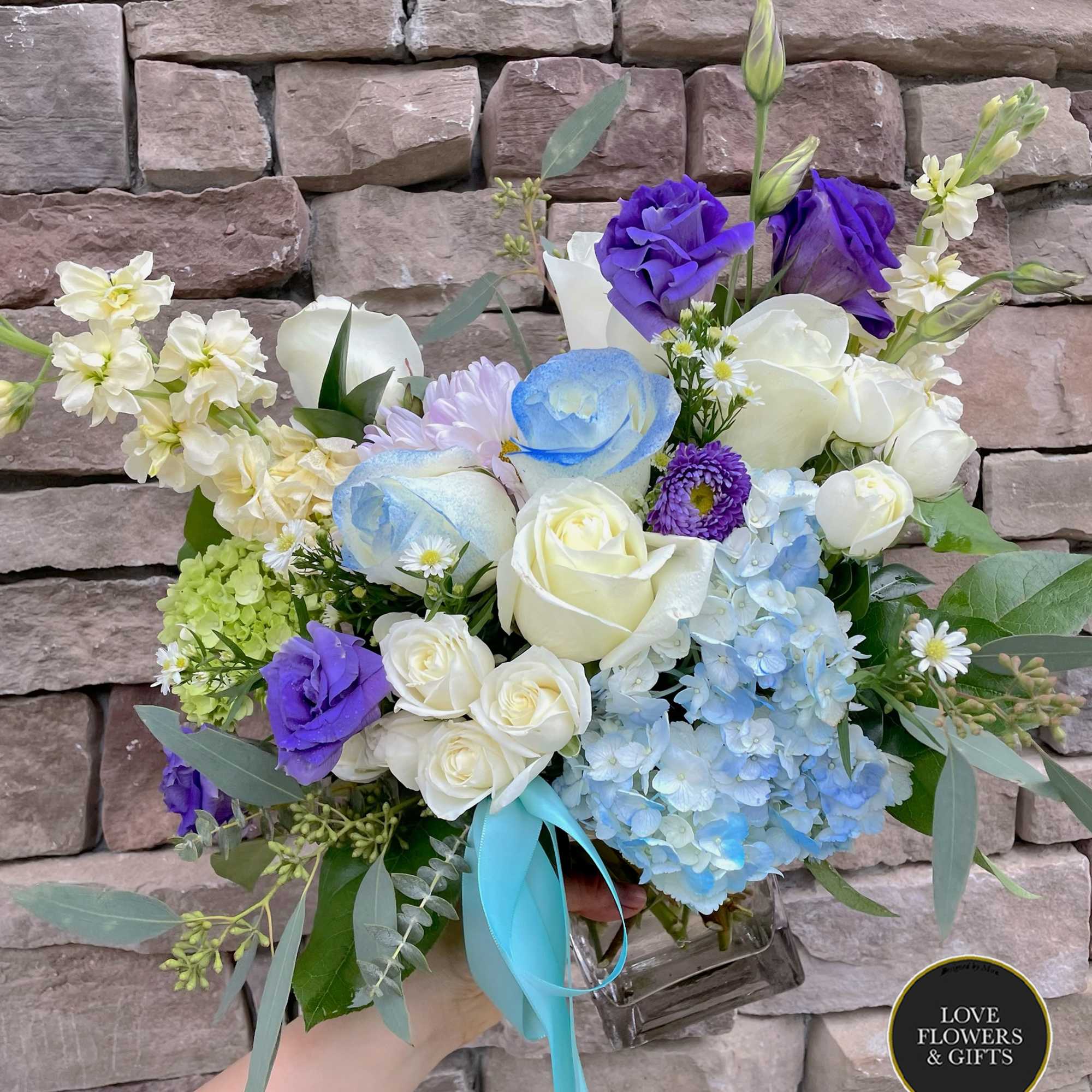 Handheld bouquet of white and blue roses with purple lisianthus and blue hydrangeas in a clear vase