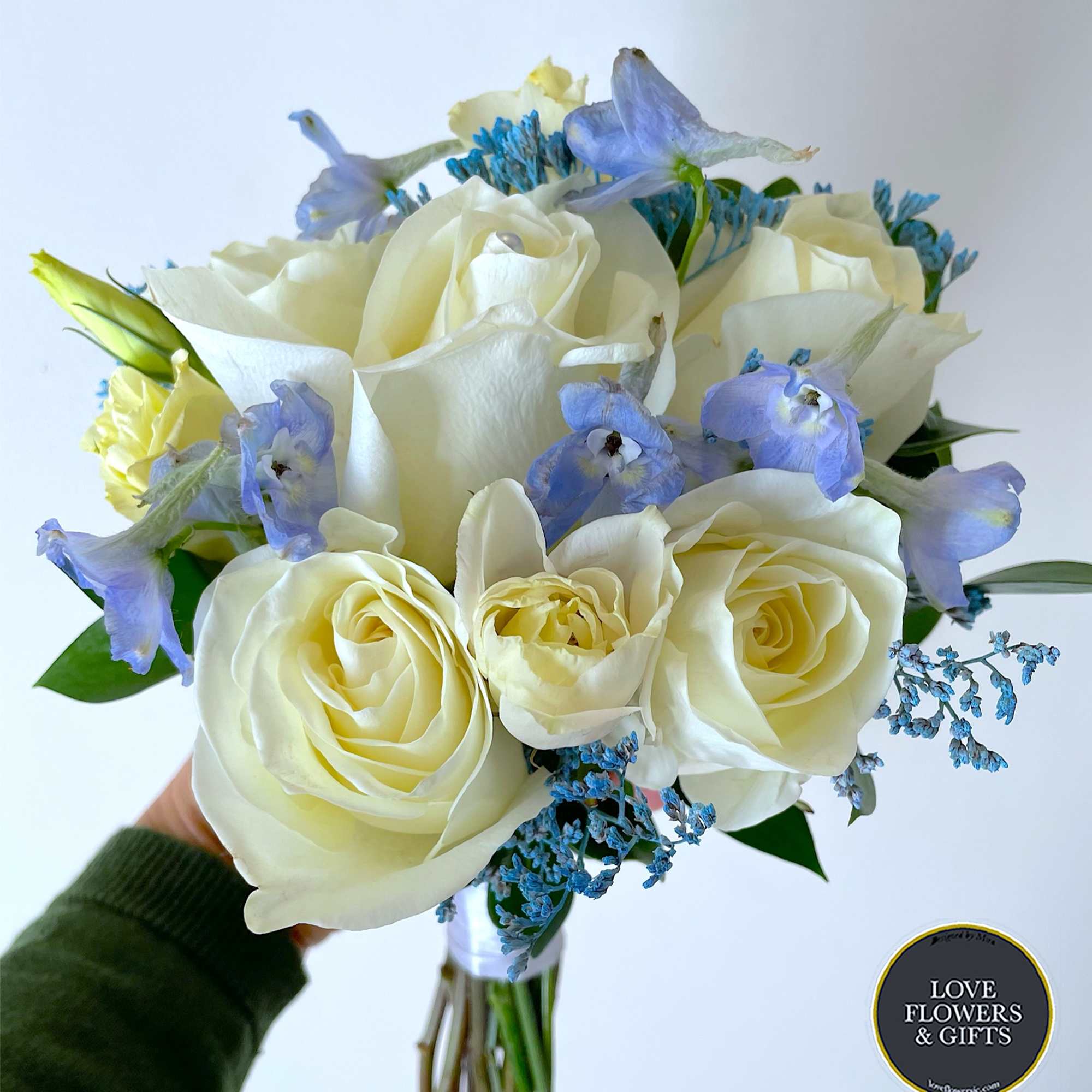Hand-tied bouquet of ivory roses with light blue flowers held against a white background