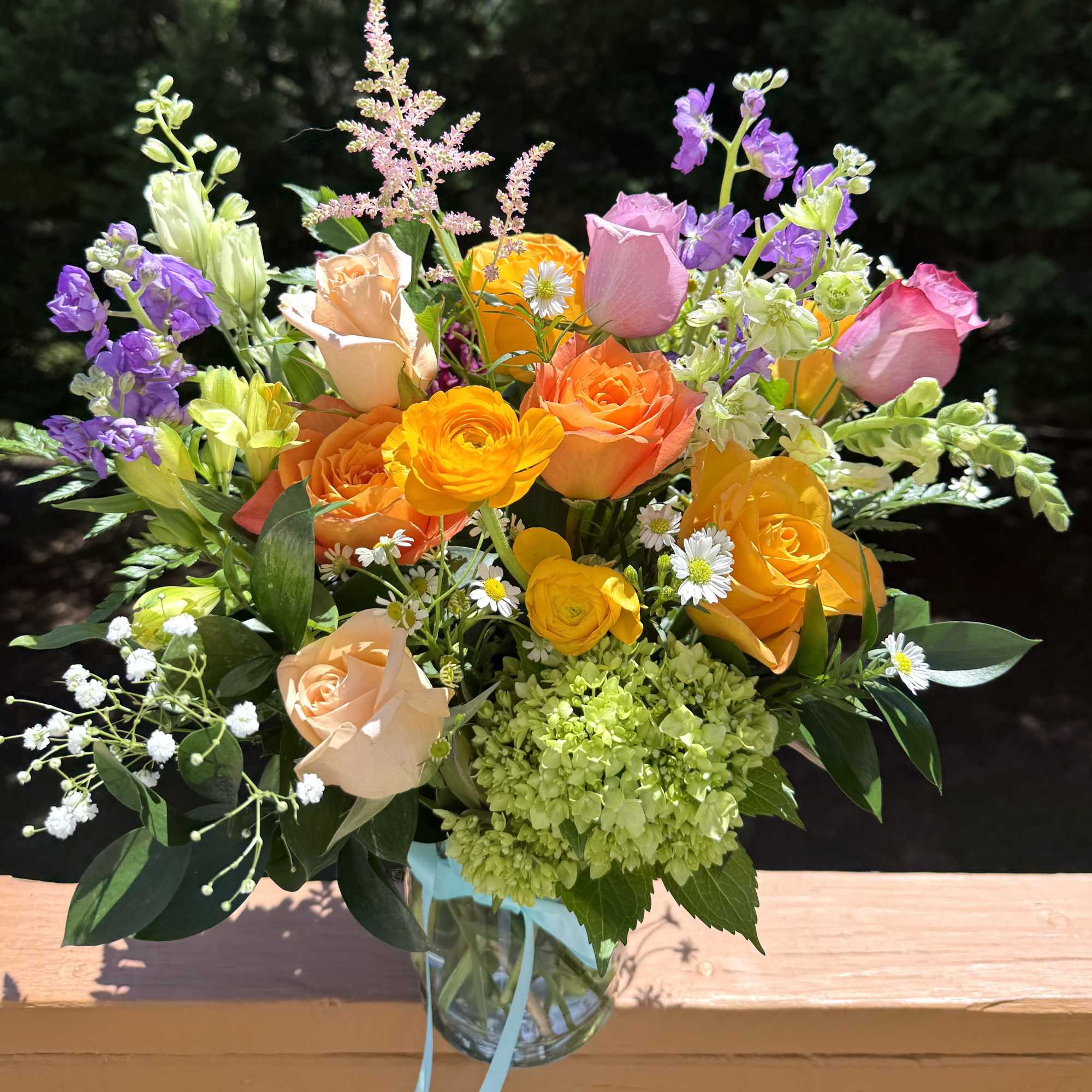 Mixed bouquet of orange and peach roses, yellow blooms, and purple accents in a clear glass vase with ribbon.