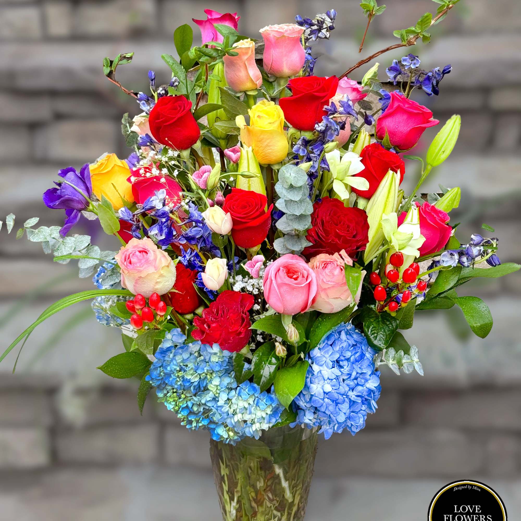 Tall mixed bouquet of multicolored roses, lilies, irises, and blue hydrangeas in a glass vase