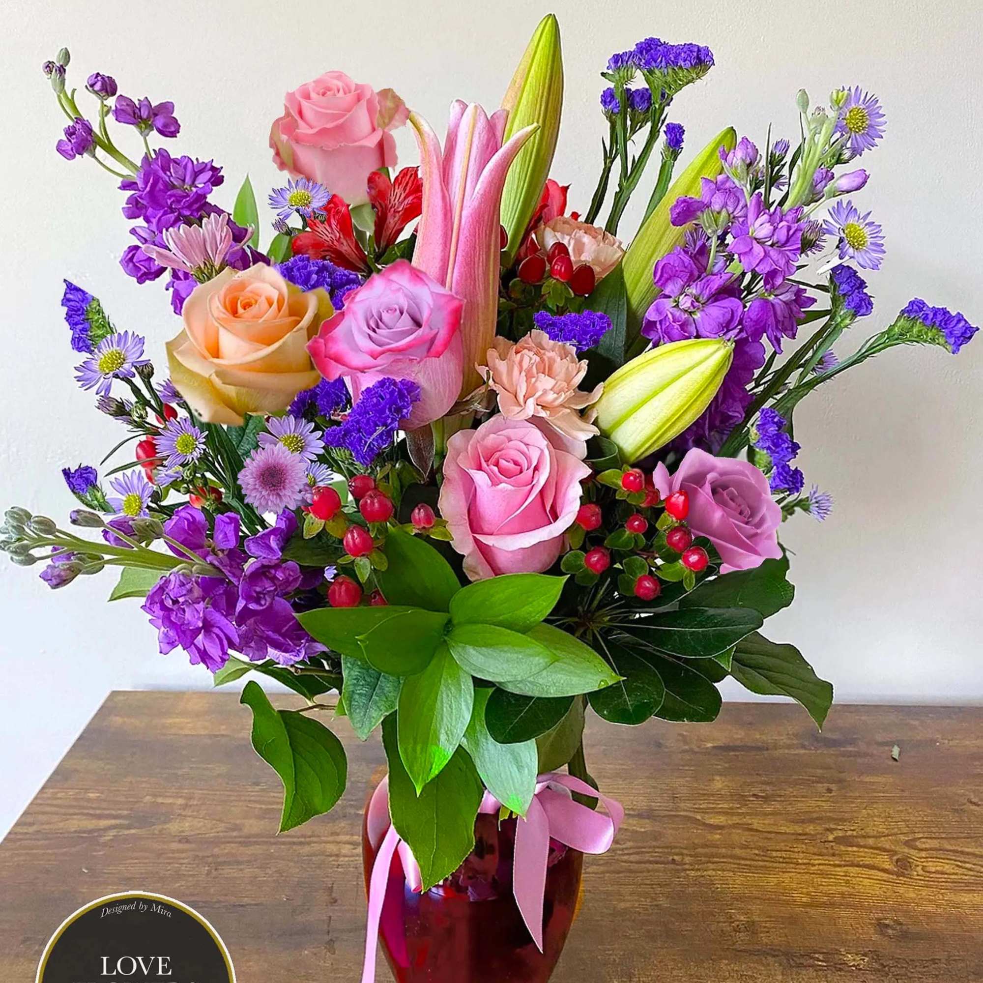 Mixed bouquet of pink roses, lilies, and purple flowers in a red glass vase with pink ribbon.
