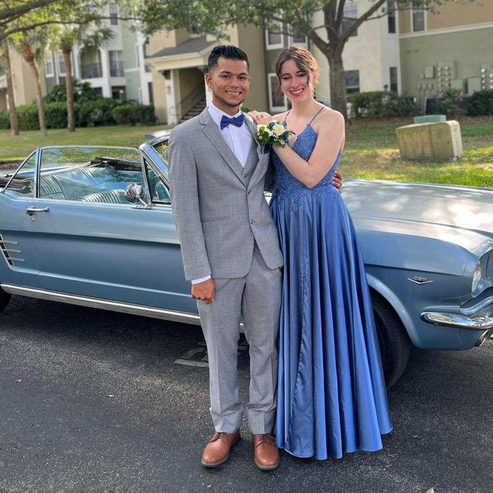 Young couple in formal attire stands by a blue convertible, with a white floral wrist corsage.