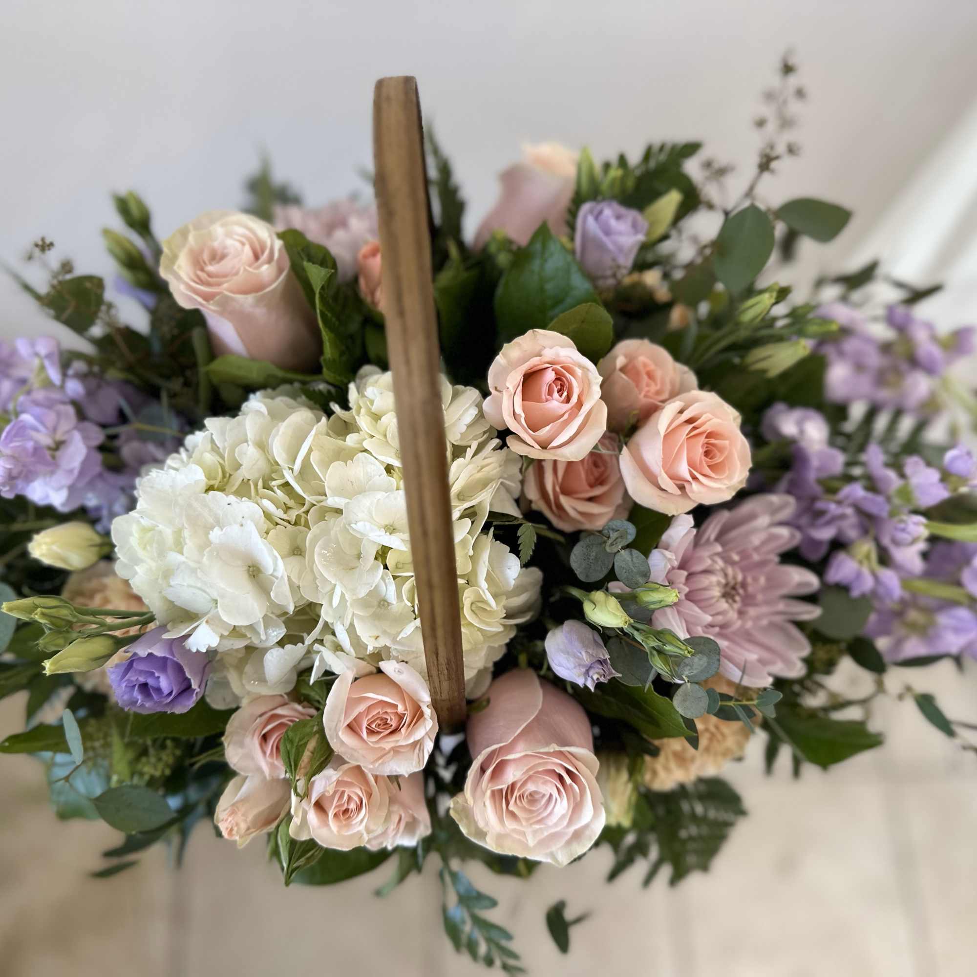 Pastel arrangement of peach roses, white hydrangeas, and lavender blooms in a wooden handled basket.