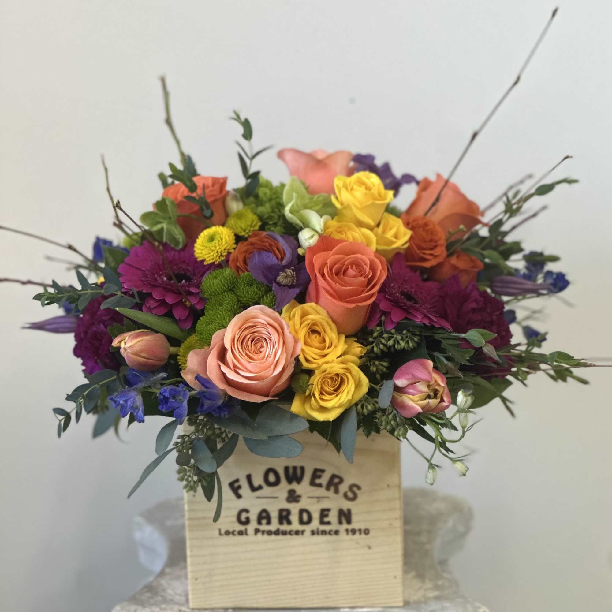 Bright mixed arrangement of roses, gerberas, tulips and mums in a wooden box on a pedestal.