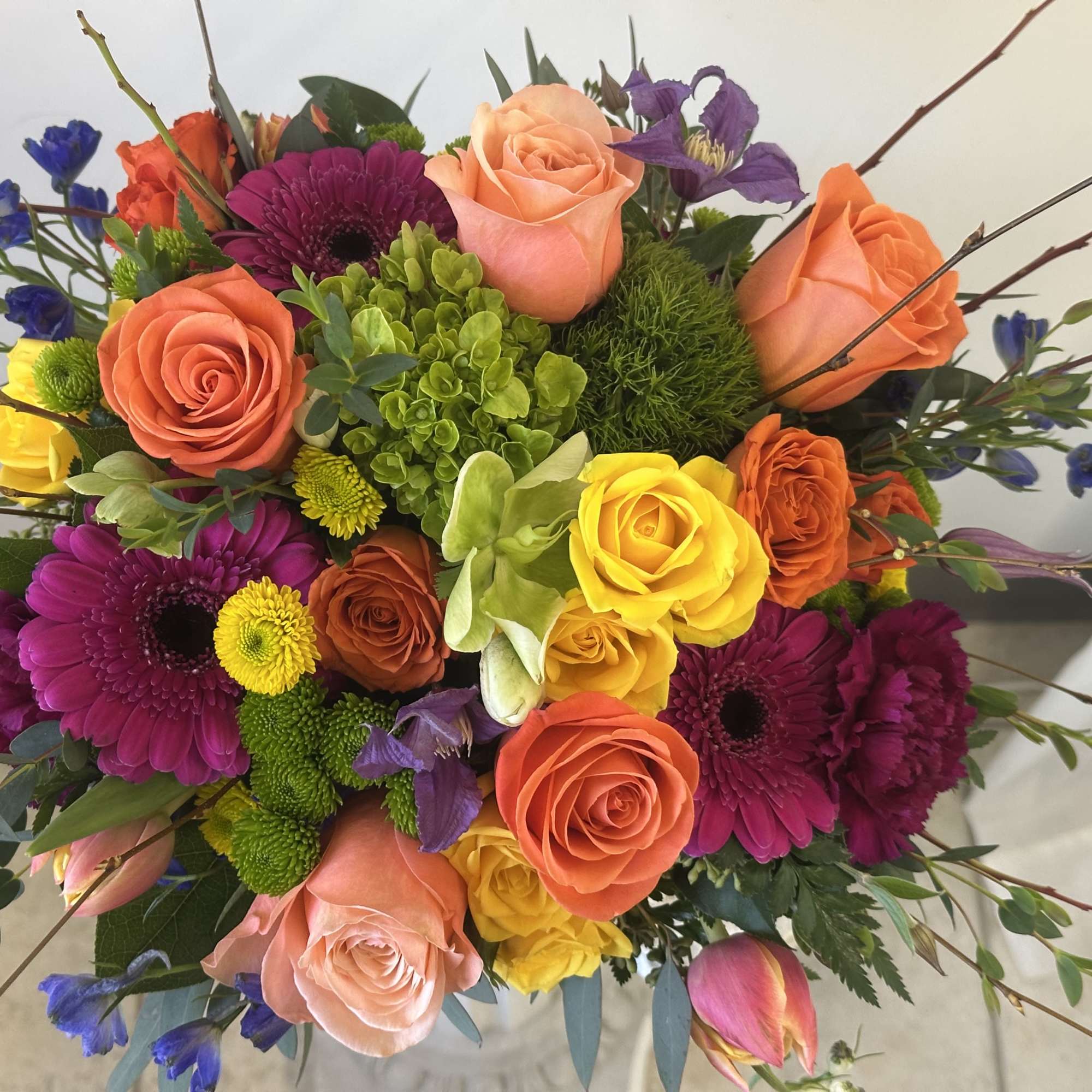 Vibrant mixed bouquet with orange and yellow roses, magenta gerberas, green blooms and purple accents in a glass vase
