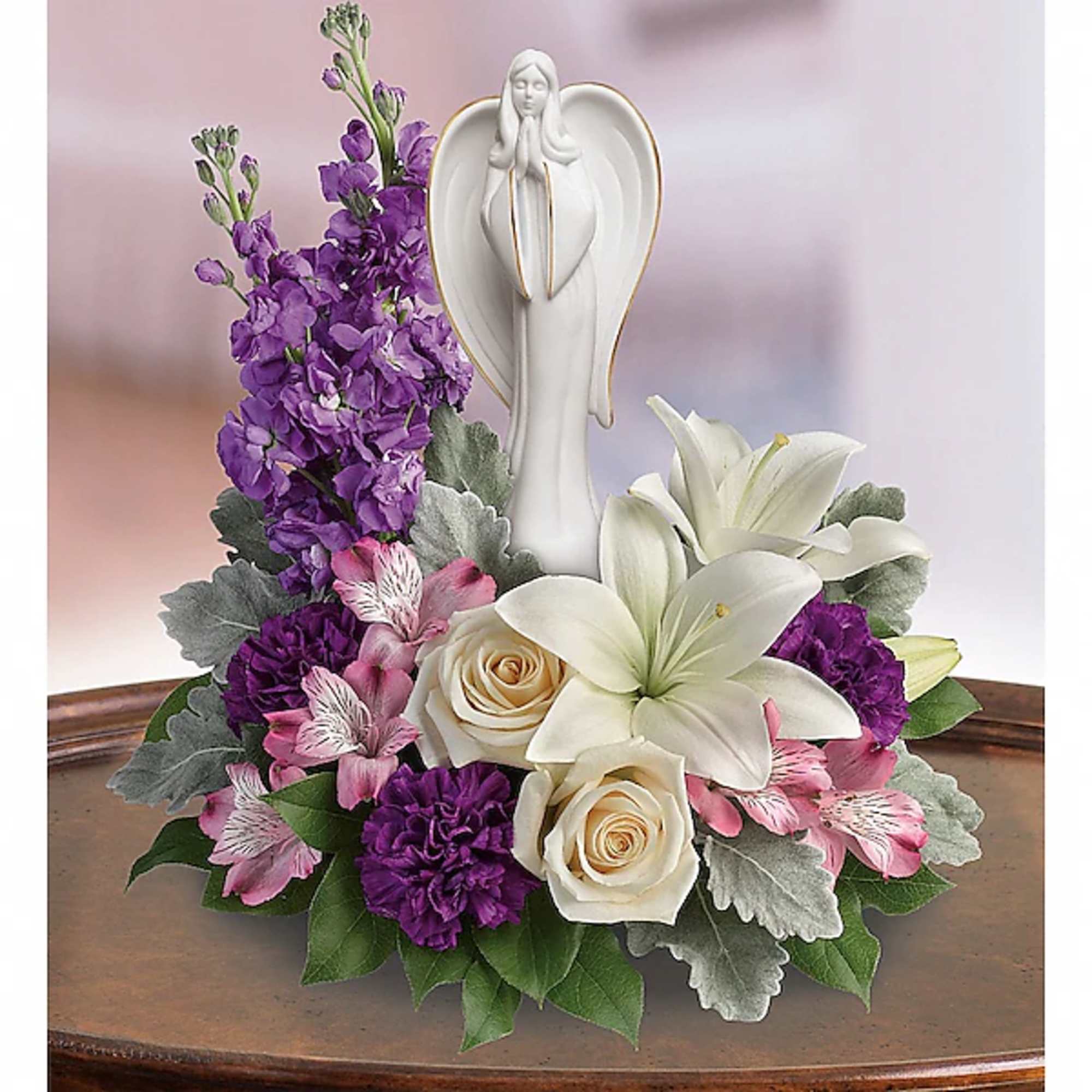 Purple and pink floral arrangement with white lilies and roses around an angel figurine