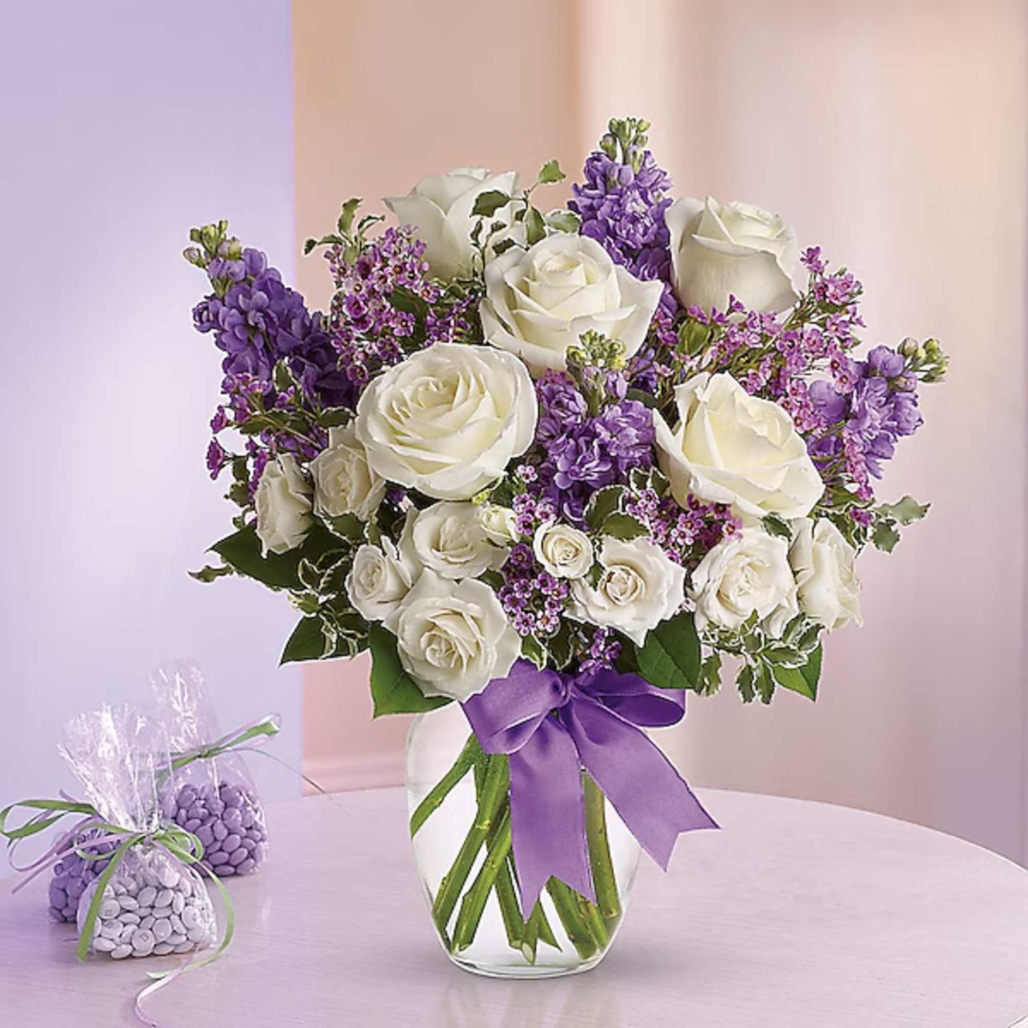 White roses and purple flowers arranged in a glass vase with a purple ribbon