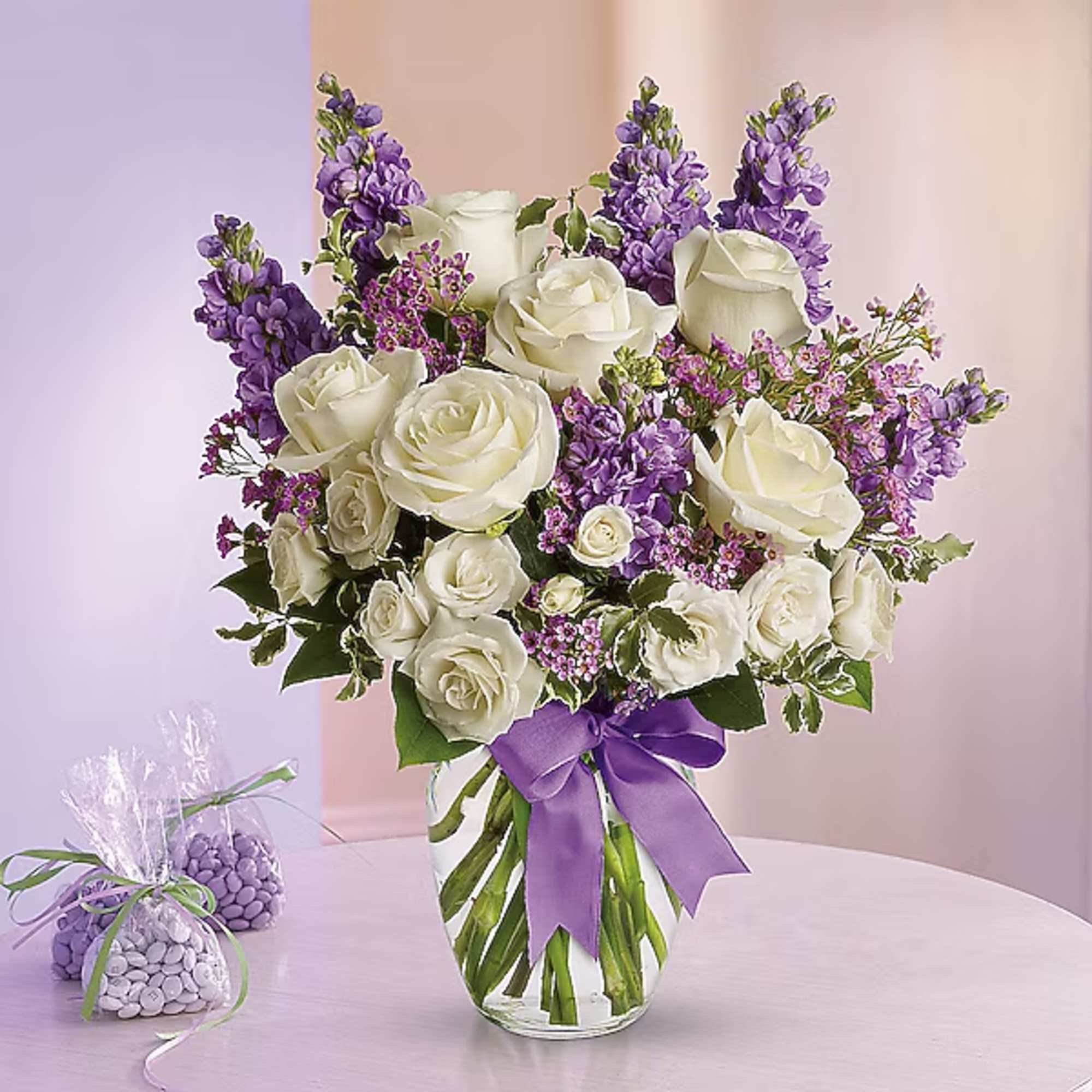 White roses and purple flowers arranged in a glass vase with a purple ribbon