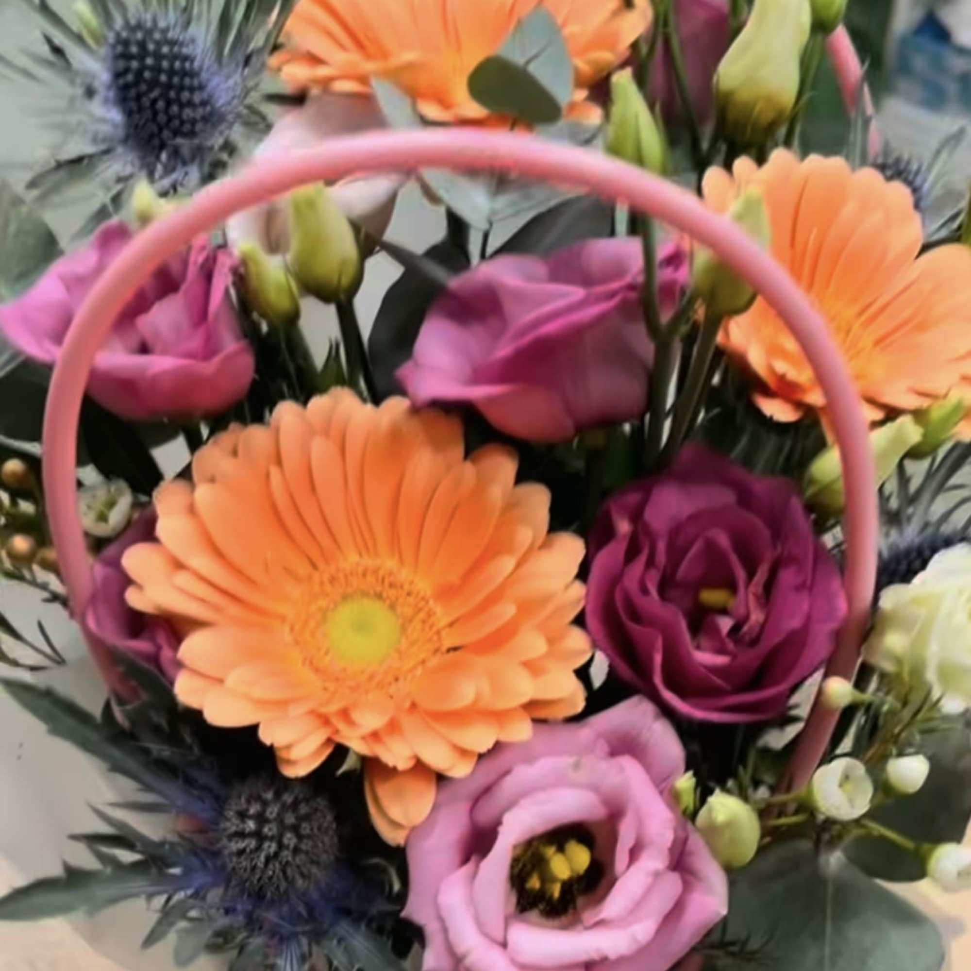Bright arrangement of orange gerbera daisies and purple lisianthus in a pink handled box