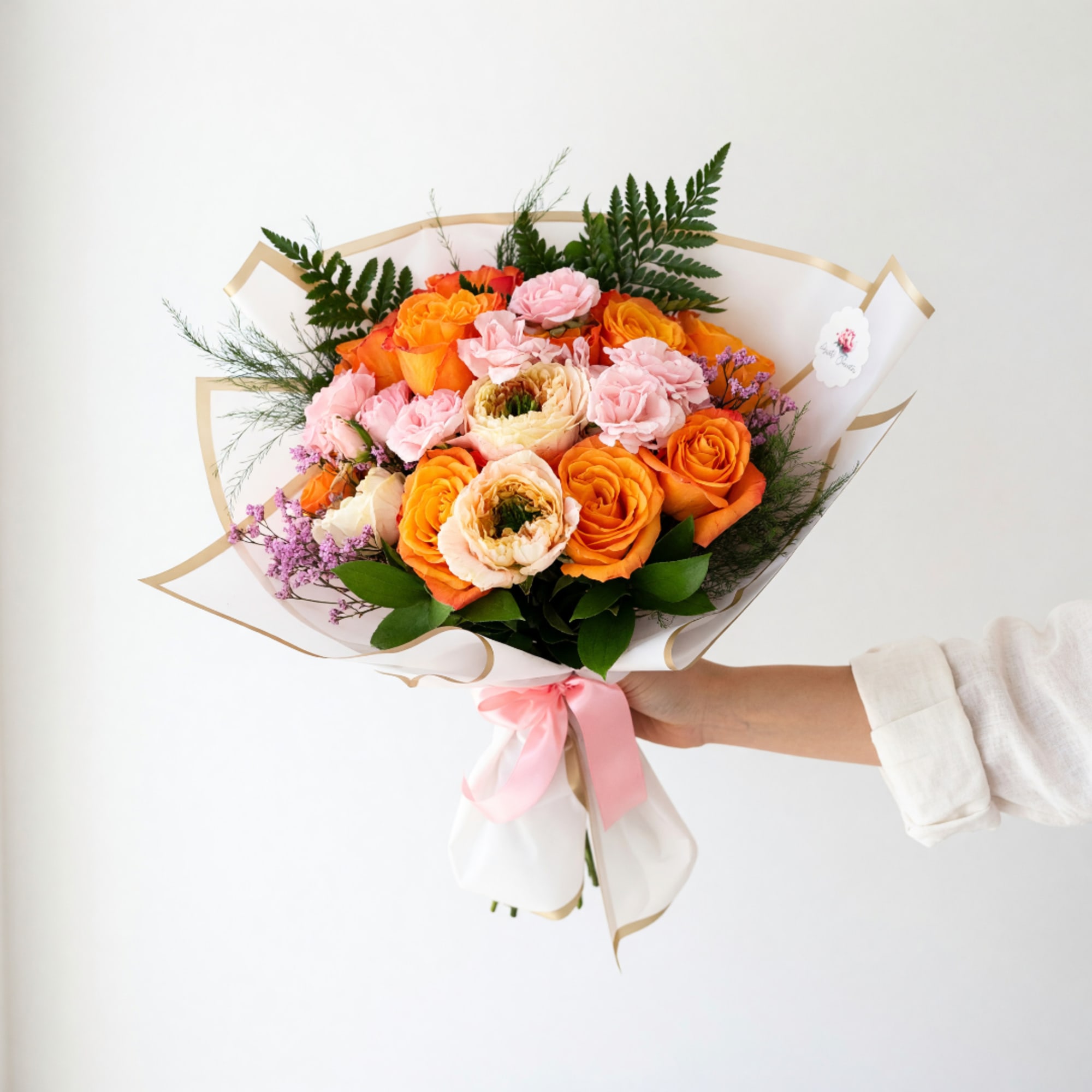 Handheld bouquet of orange and pink roses wrapped in white paper with a pink ribbon