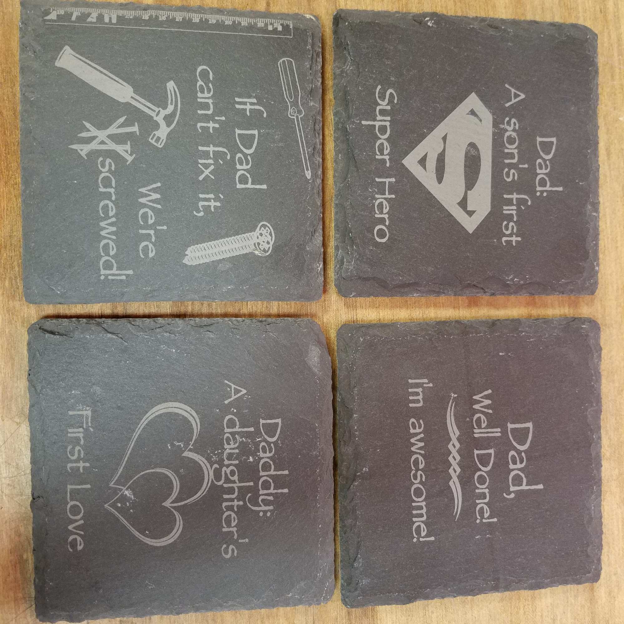 Four engraved slate coasters with father-themed sayings on a wooden surface.
