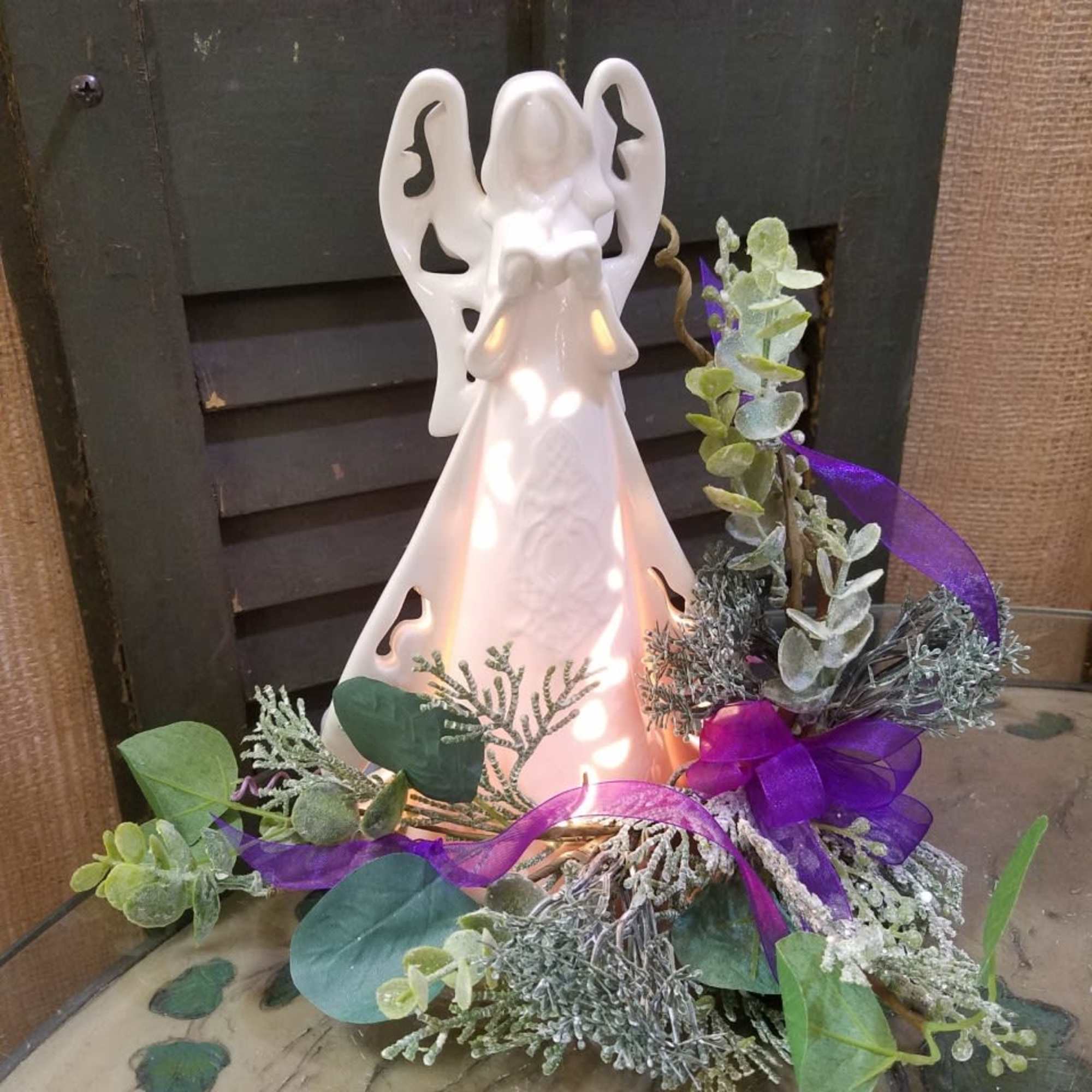 White angel figurine with purple ribbon and greenery arrangement