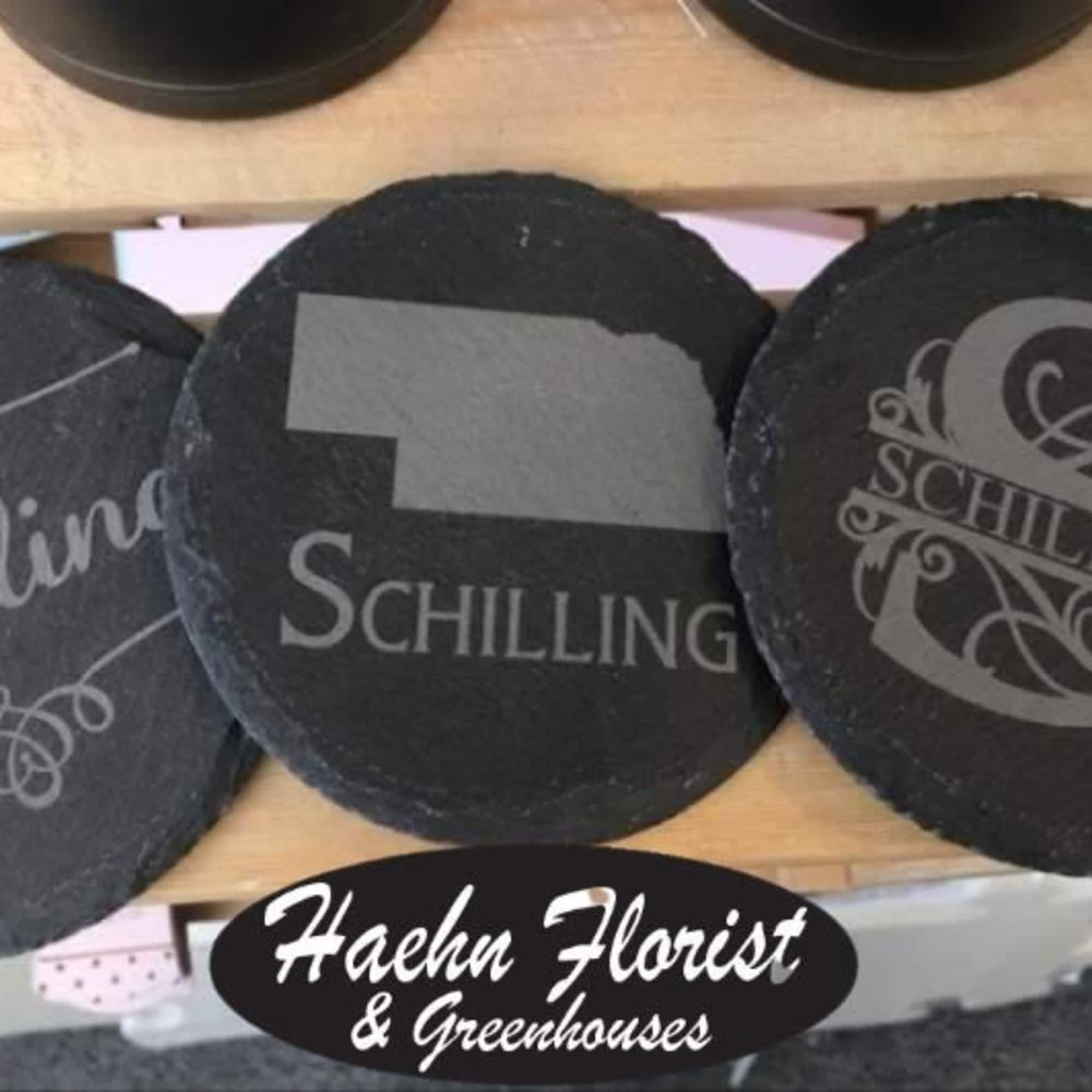 Three round black slate coasters with Schilling logos on a wooden shelf.