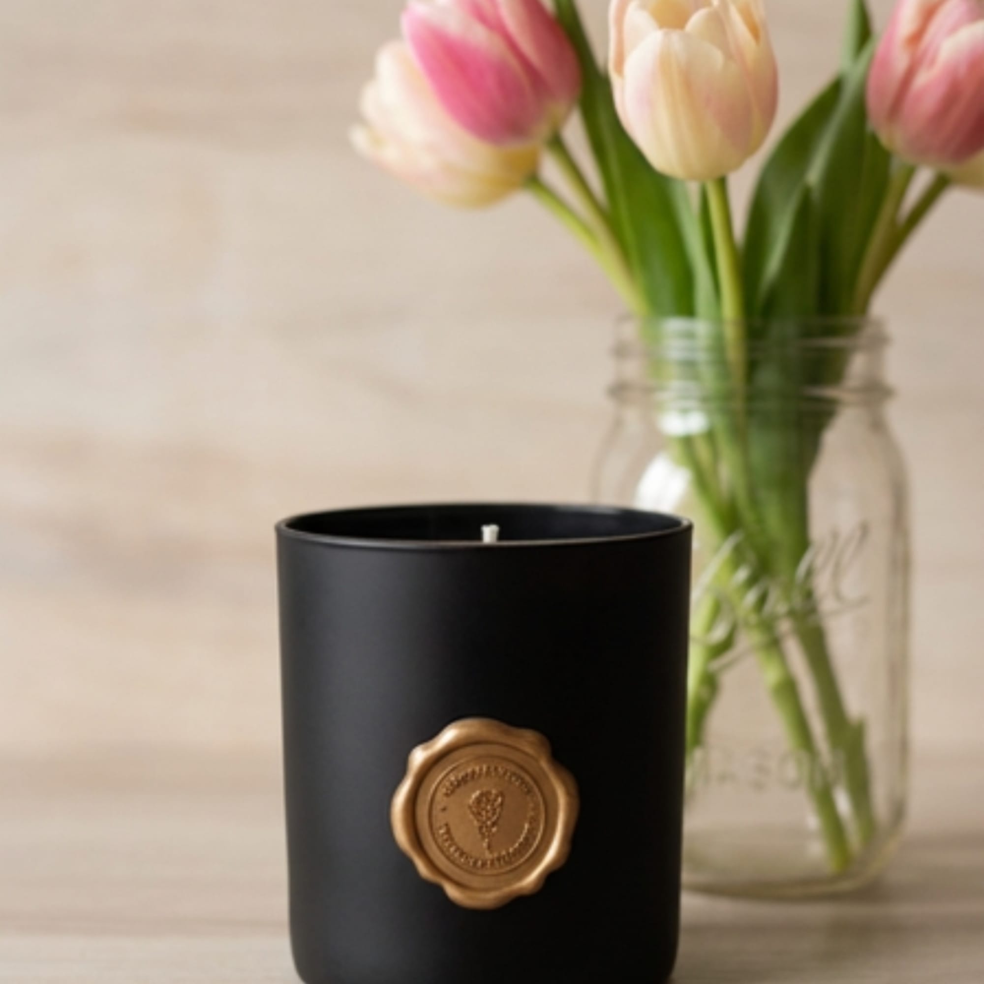 Black candle in a matte jar with pink tulips in a glass vase behind it